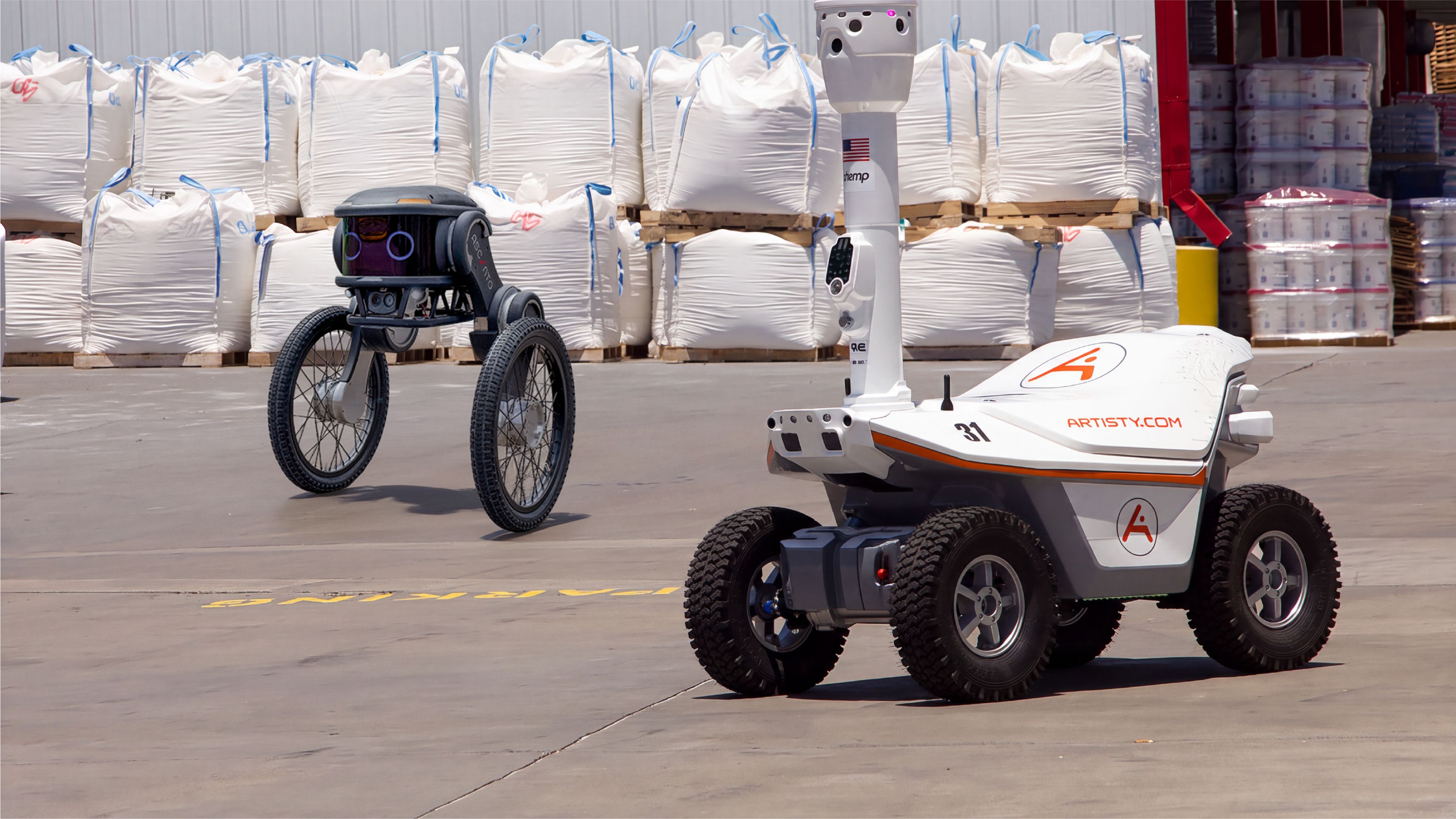 Two outdoor security patrol robots at a warehouse loading area: a compact two-wheel Ascento Guard robot in the background, and a four-wheel Artisty-branded patrol robot with a raised camera mast in the foreground."