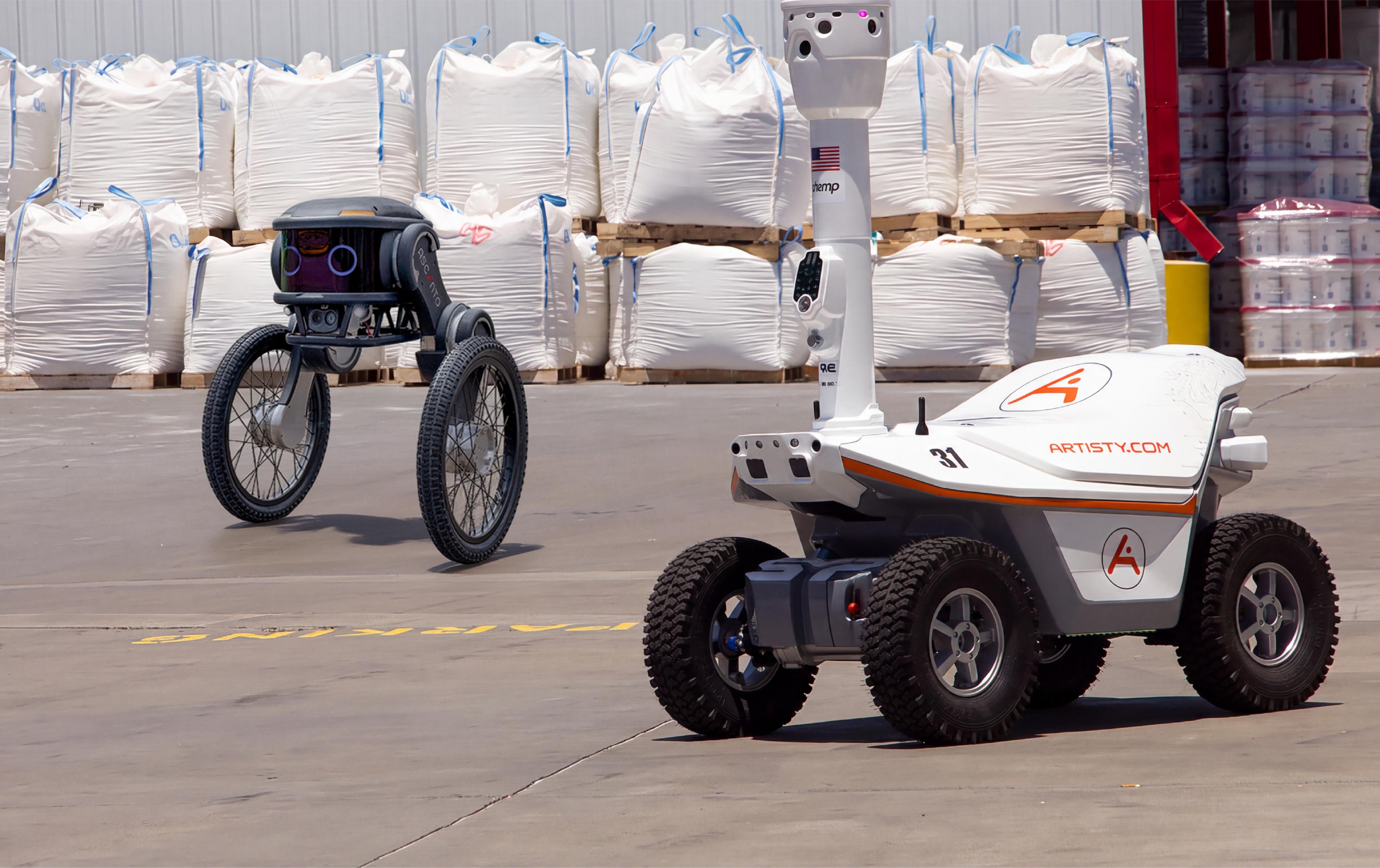 Two outdoor security patrol robots at a warehouse loading area: a compact two-wheel Ascento Guard robot in the background, and a four-wheel Artisty-branded patrol robot with a raised camera mast in the foreground."