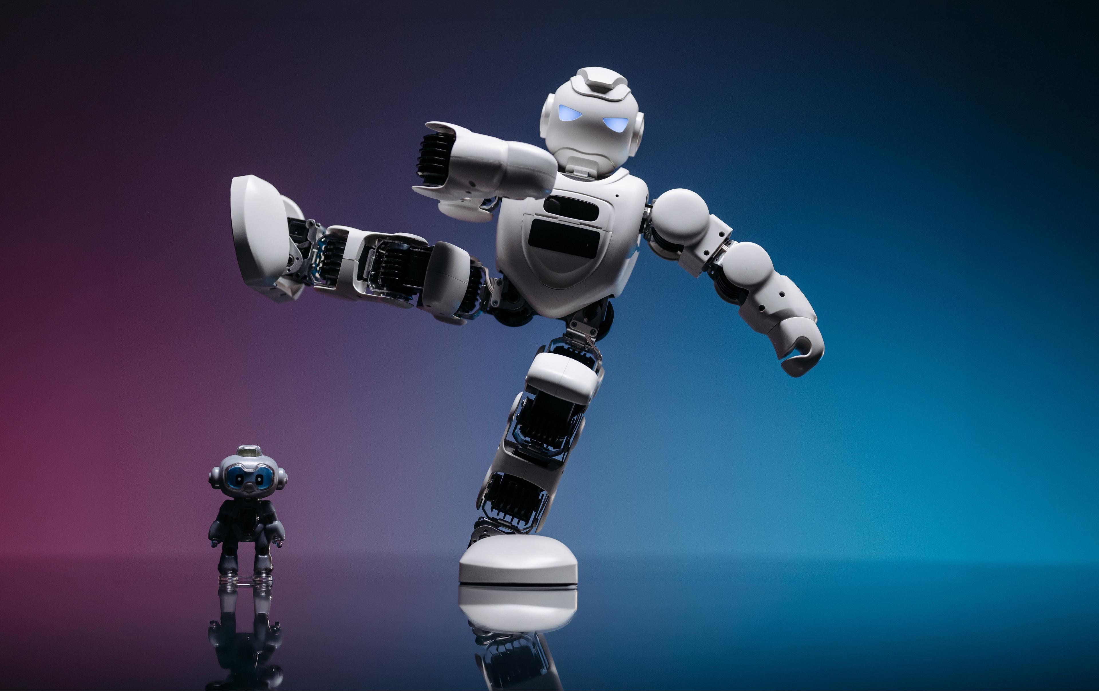 White humanoid robot balancing on one leg in a high kick next to a small black humanoid on a reflective studio floor
