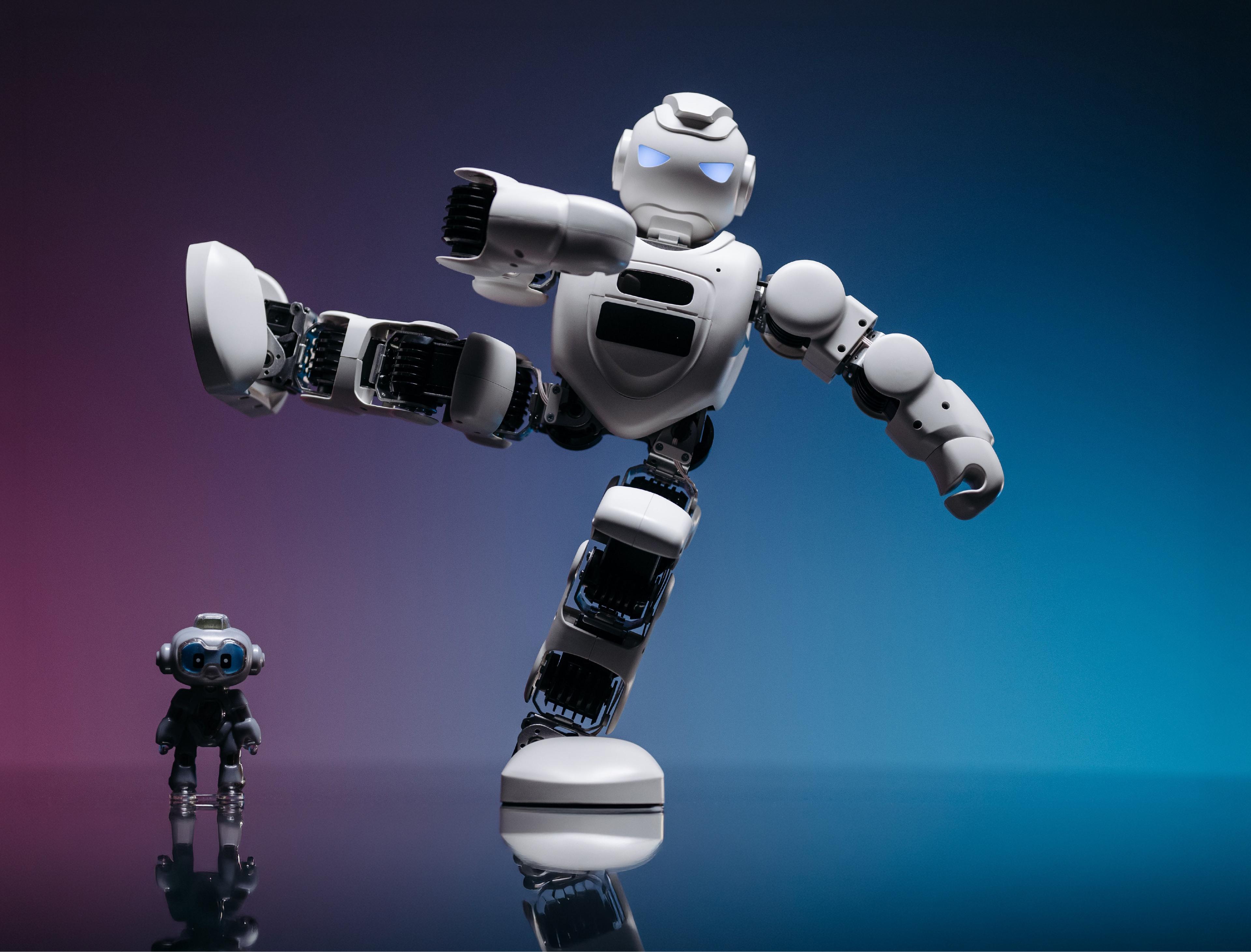 White humanoid robot balancing on one leg in a high kick next to a small black humanoid on a reflective studio floor