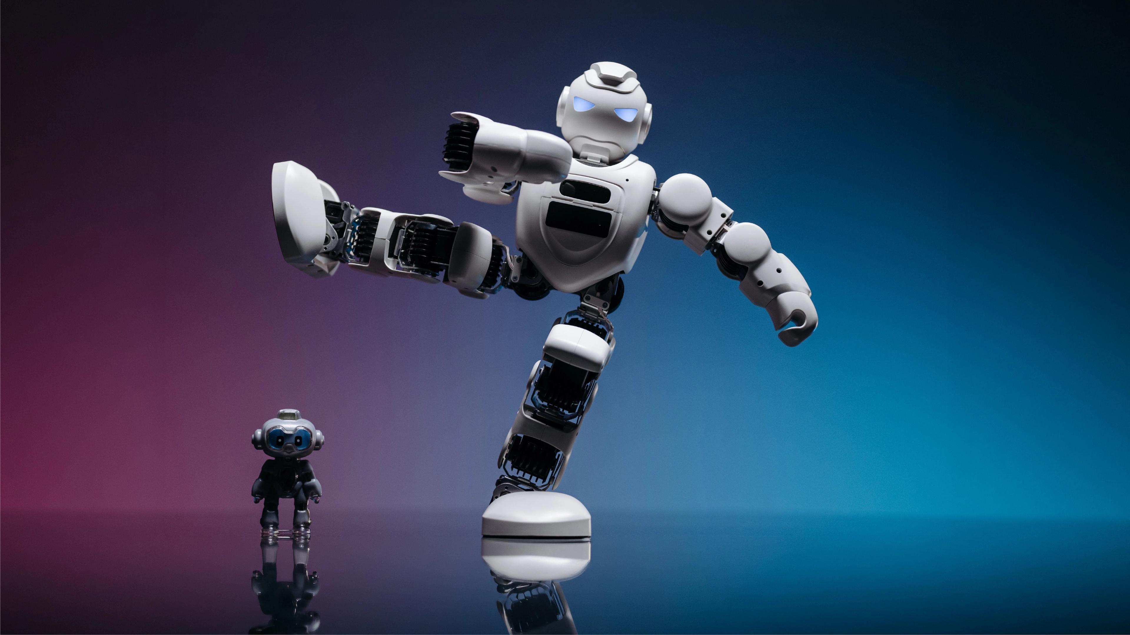 White humanoid robot balancing on one leg in a high kick next to a small black humanoid on a reflective studio floor