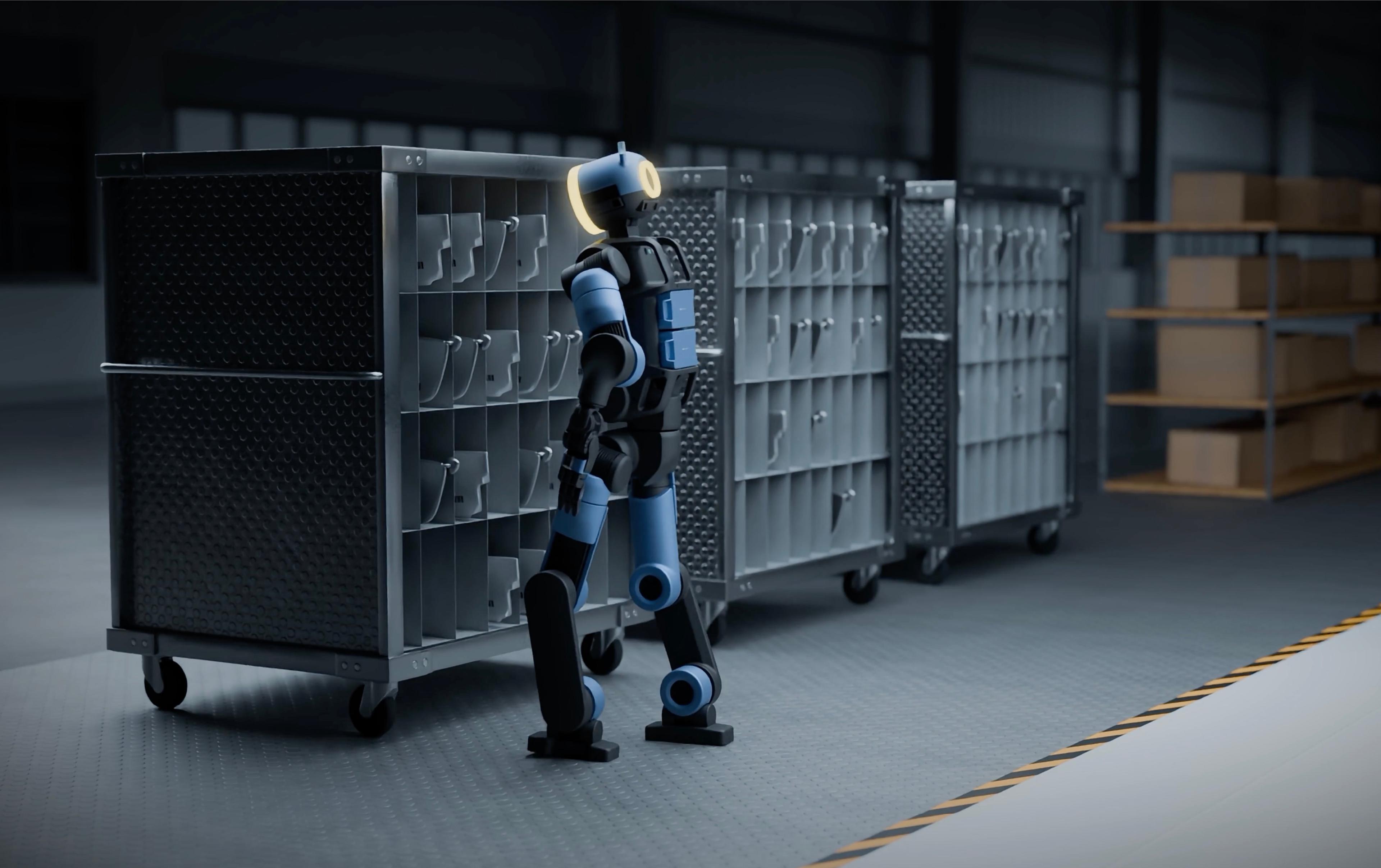 A bipedal humanoid robot stands in profile facing a line of metal parts carts in a dimly lit industrial warehouse.