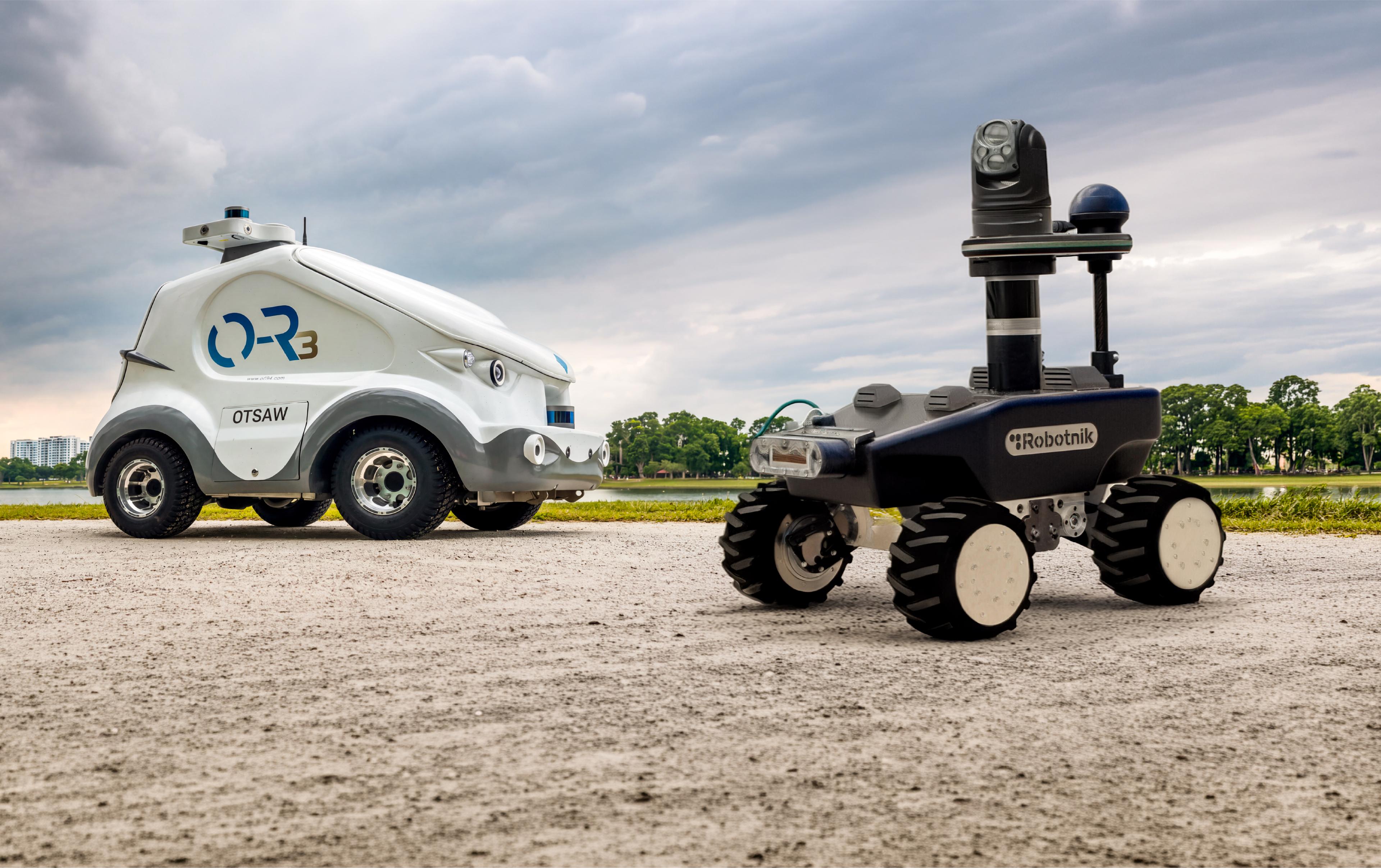 Two outdoor security patrol robots on a gravel path beside a park lake: OTSAW's compact white car-like O-R3 robot on the left, and Robotnik's dark RB-WATCHER with a raised PTZ camera and sensor mast on the right