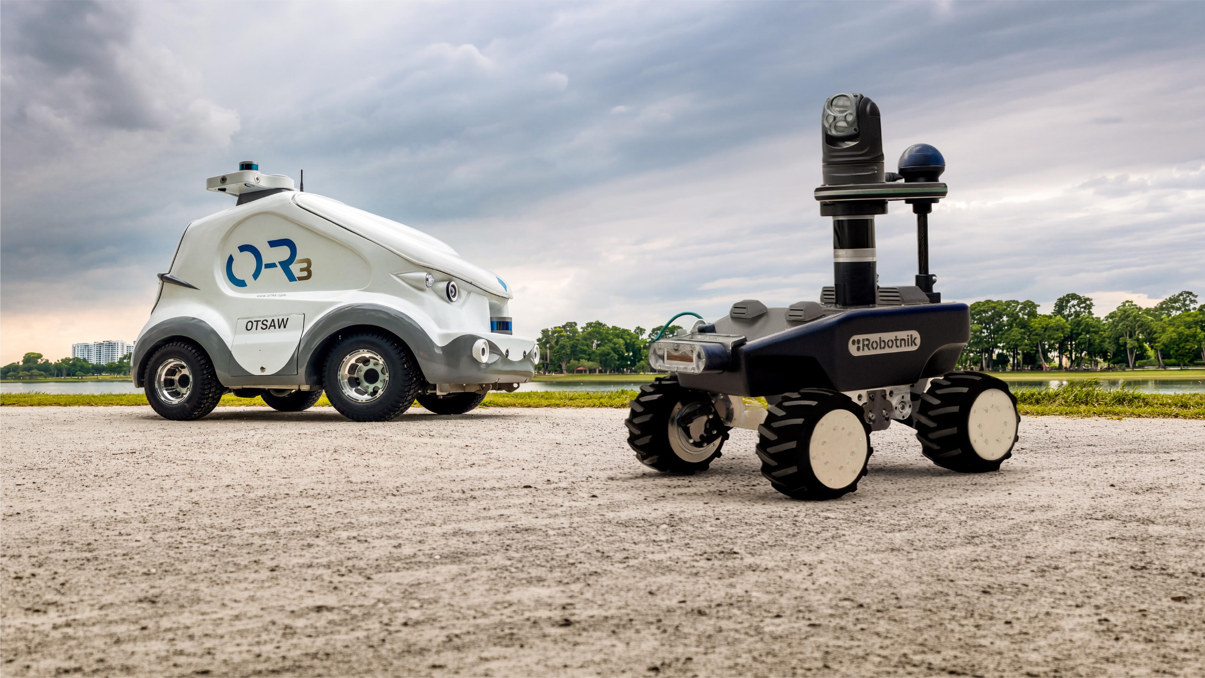 Two outdoor security patrol robots on a gravel path beside a park lake: OTSAW's compact white car-like O-R3 robot on the left, and Robotnik's dark RB-WATCHER with a raised PTZ camera and sensor mast on the right