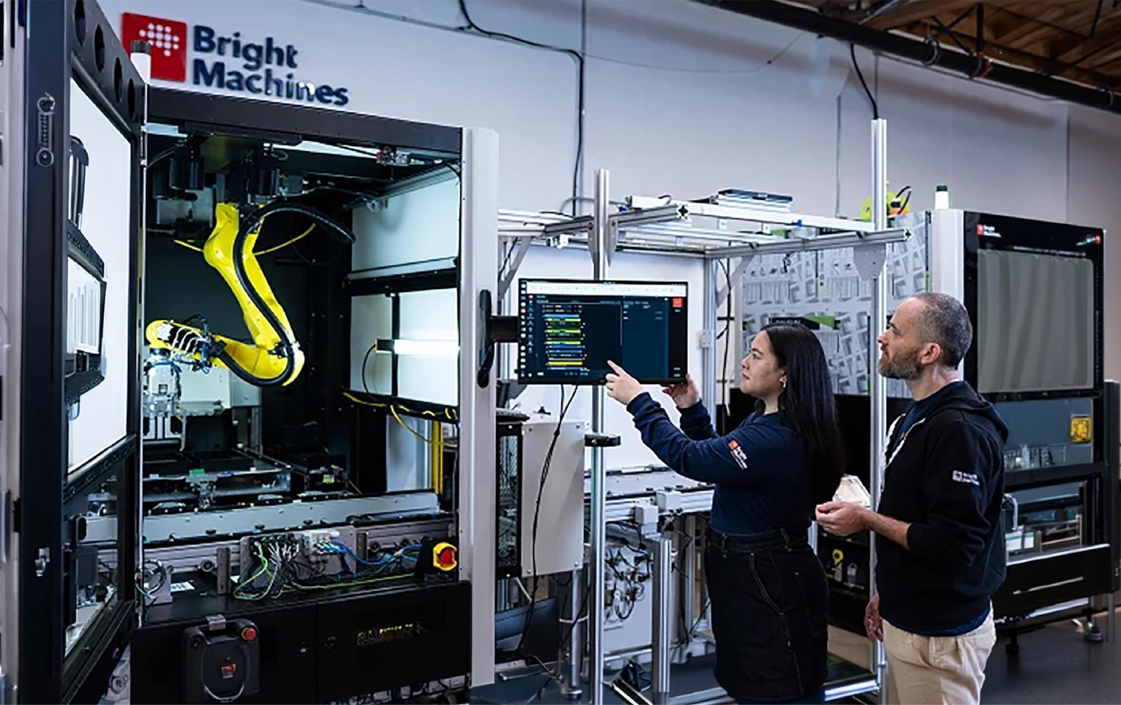 Bright Machines employees review a computer screen beside an automated manufacturing system with an industrial robotic arm.