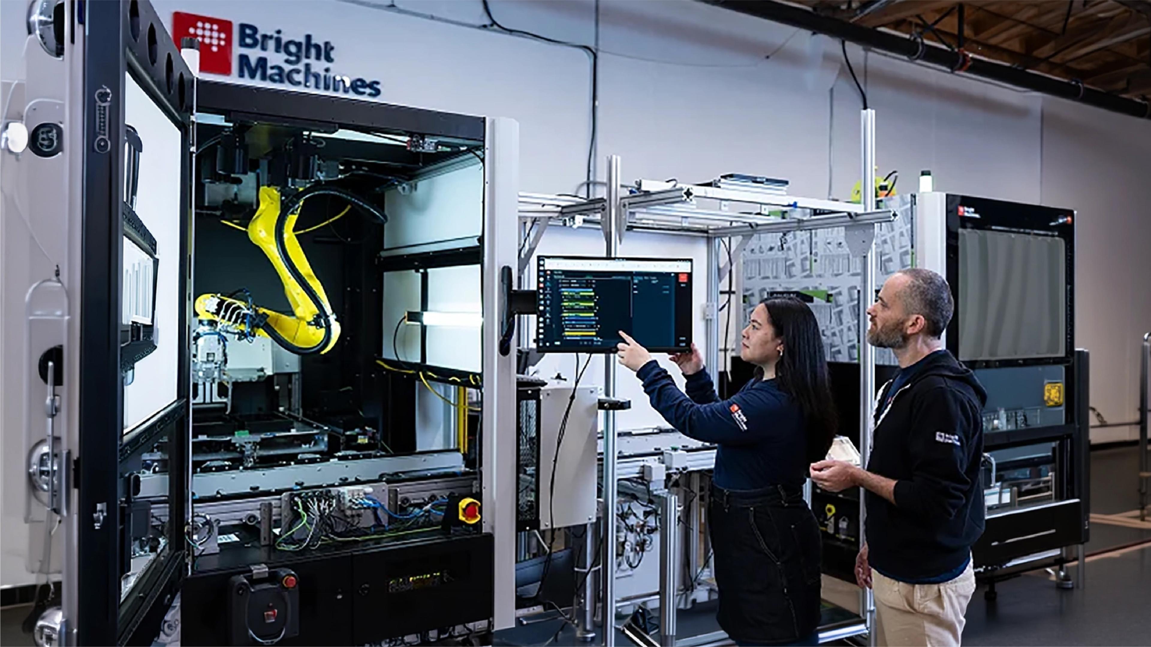 Bright Machines employees review a computer screen beside an automated manufacturing system with an industrial robotic arm.