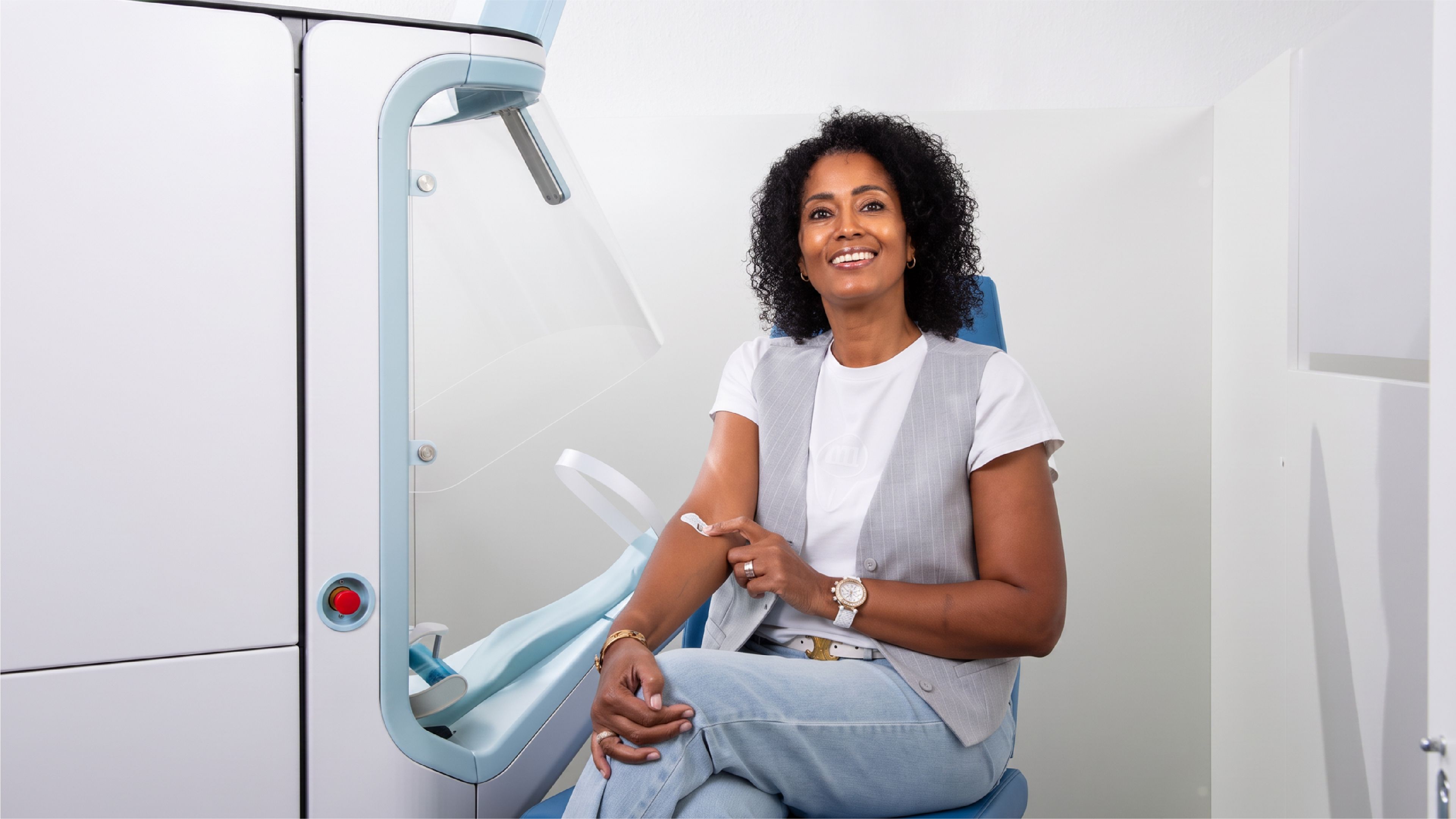 A smiling woman sits in a chair beside Aletta, an automated blood-draw device, with a small bandage on the inside of her arm after using the system.