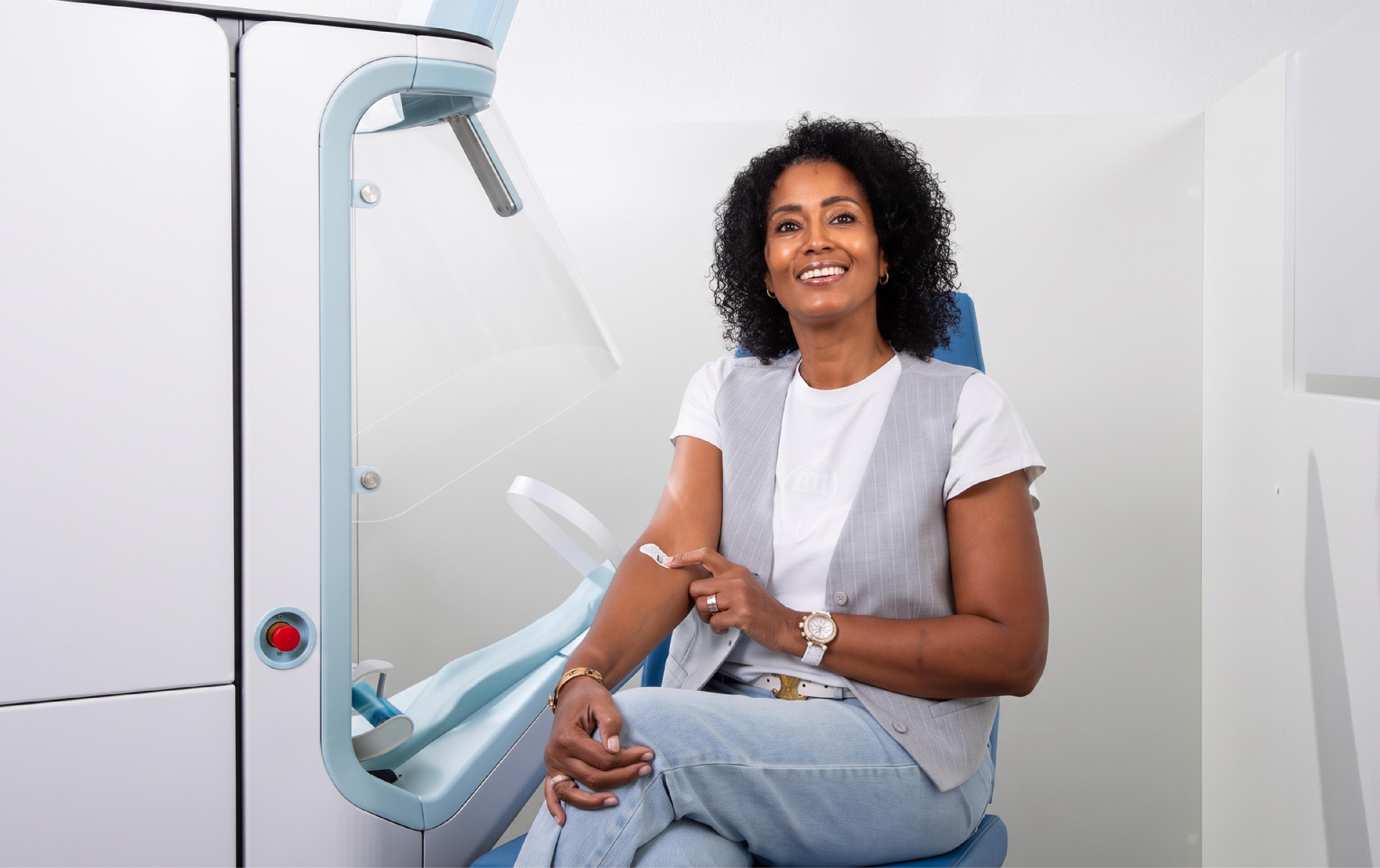 A smiling woman sits in a chair beside Aletta, an automated blood-draw device, with a small bandage on the inside of her arm after using the system.