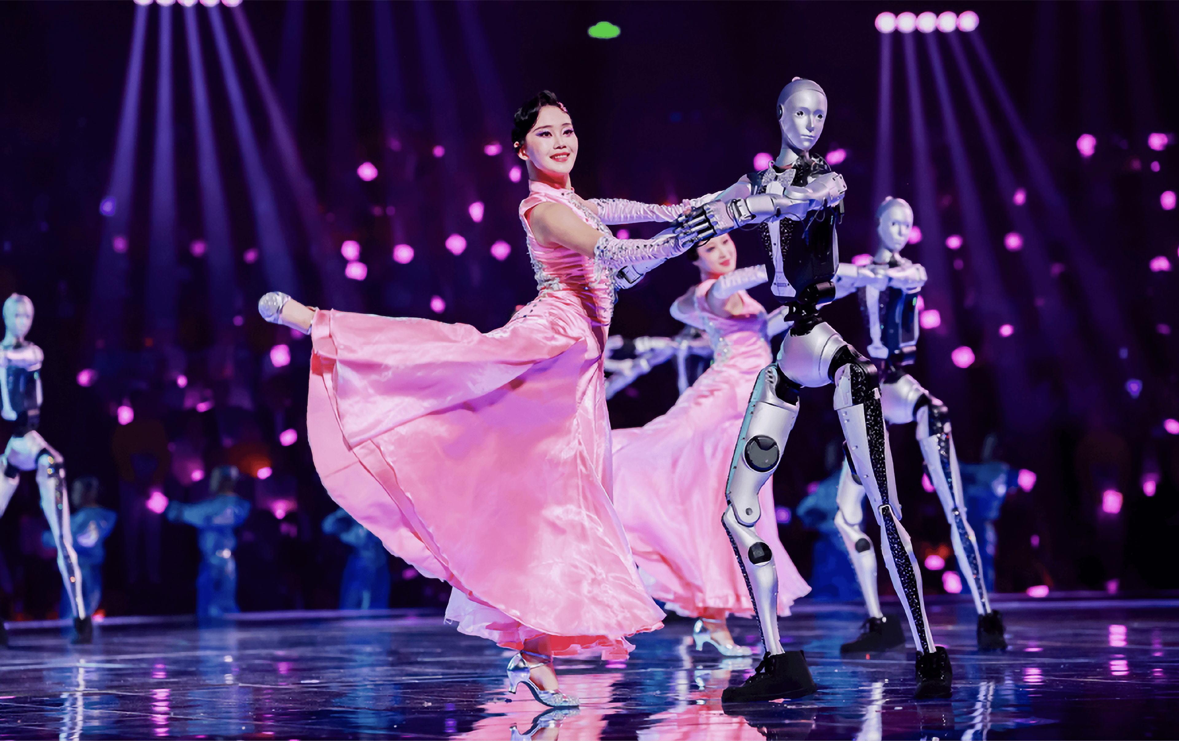 A woman in a pink dress dances joyfully with two humanoid robots in a lively setting.