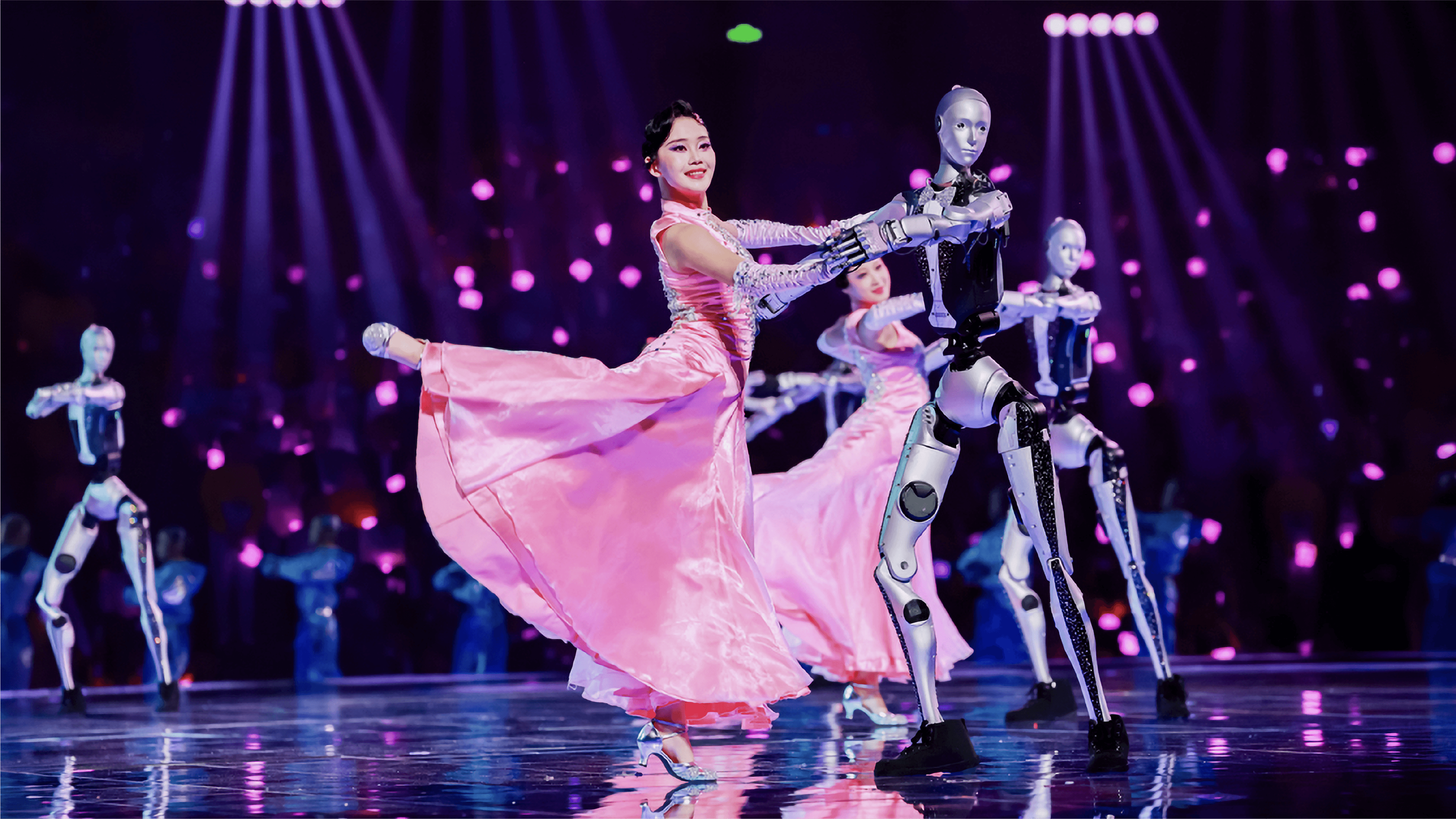 A woman in a pink dress dances joyfully with two humanoid robots in a lively setting.