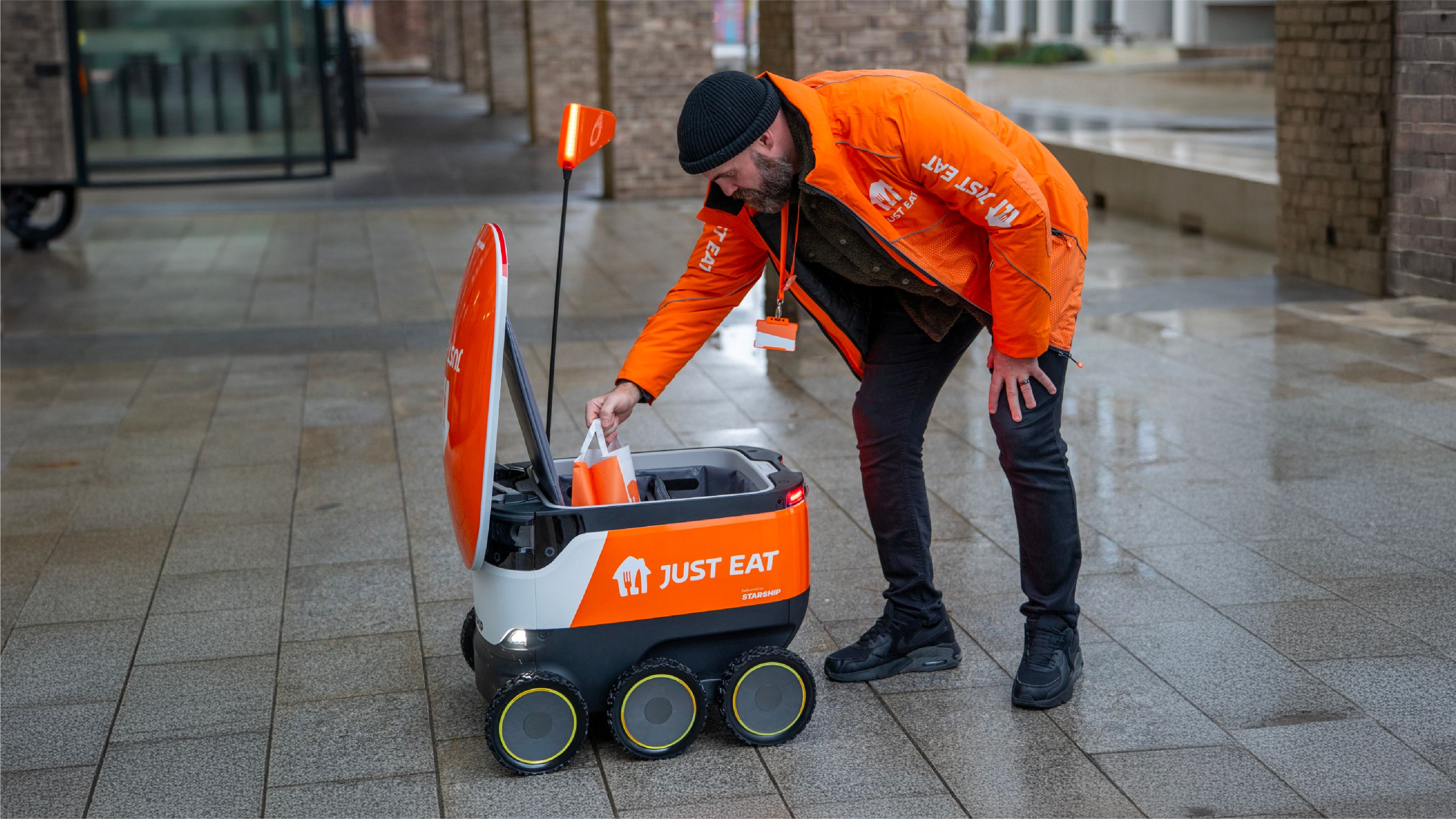 A Just Eat worker loads an order into a Starship delivery robot on a wet sidewalk.