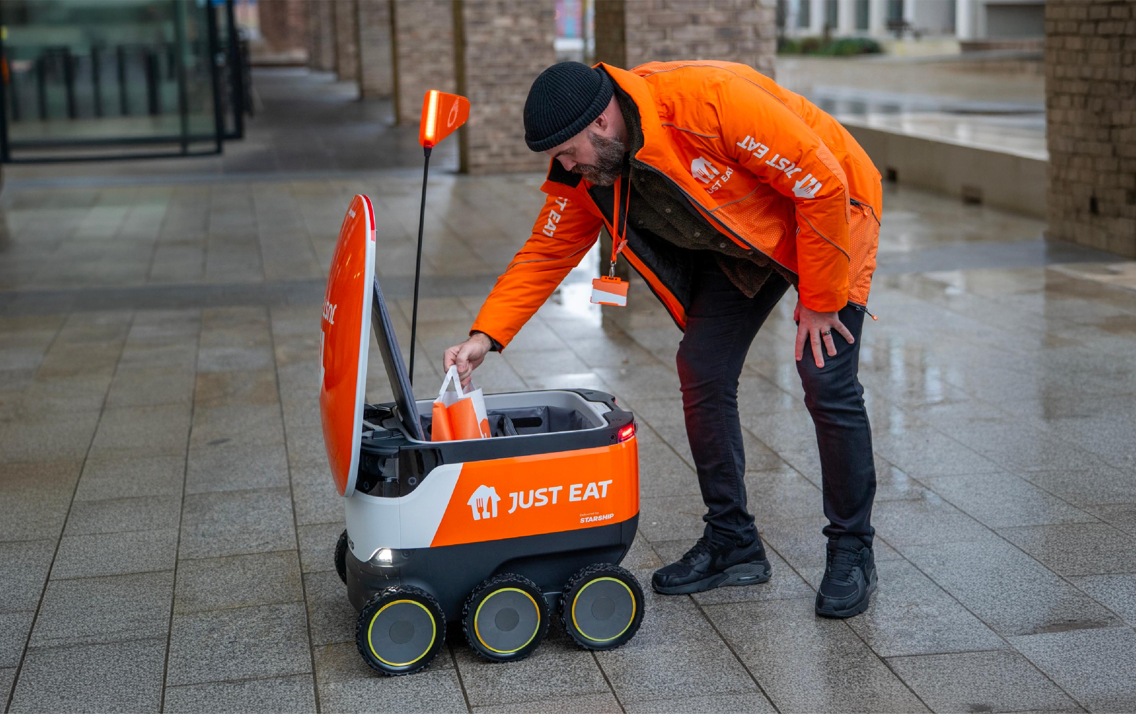 A Just Eat worker loads an order into a Starship delivery robot on a wet sidewalk.