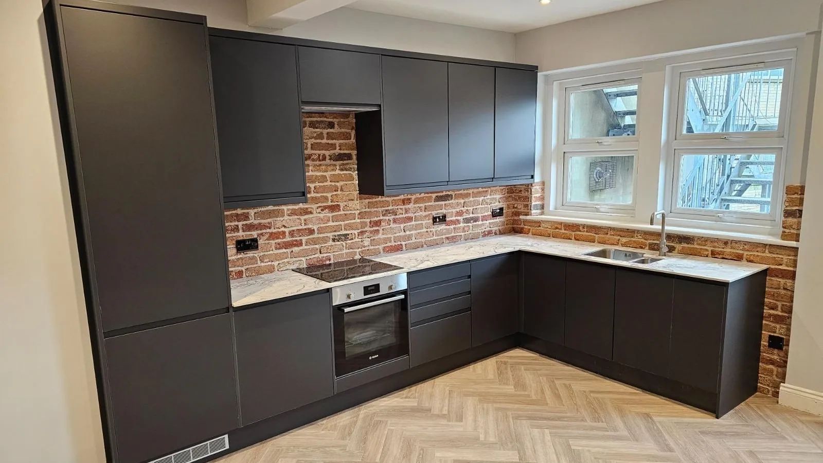Wide view of a charcoal kitchen with exposed brick, marble worktop and herringbone floor