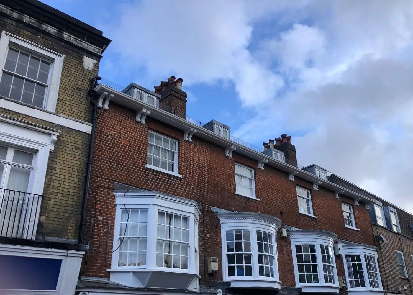 Row of brick terraced buildings with renewed white bay sash windows and chimney stacks against a cloudy sky