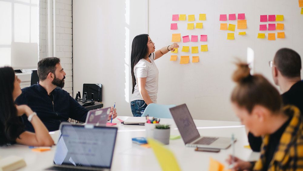 A woman is at the front of the room leading a discussion around her team's kanban board.