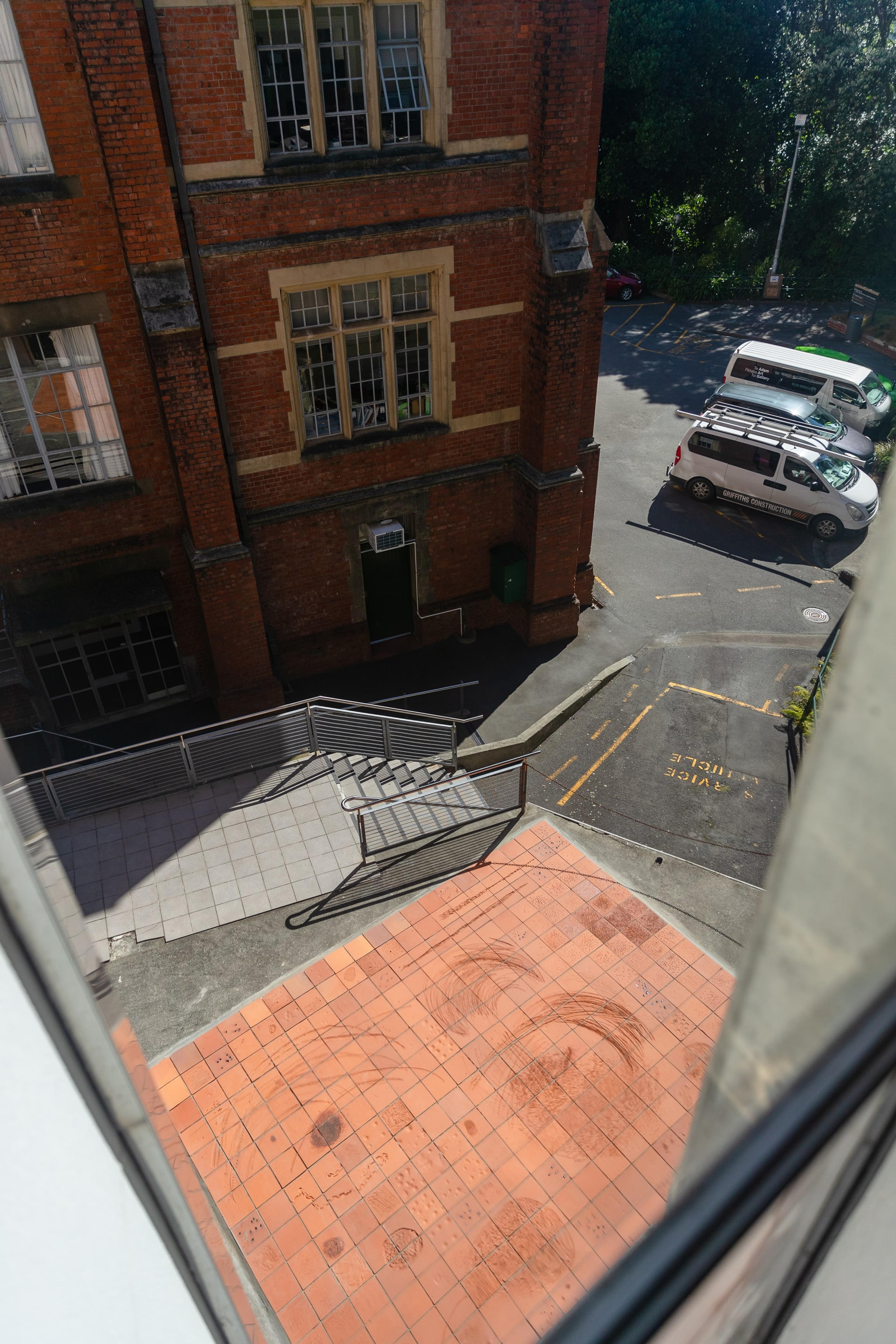 A photograph of the view looking down through a window of the gallery, at a carpark and flat tiled area covered in terracotta tiles with large scratches and marks.
