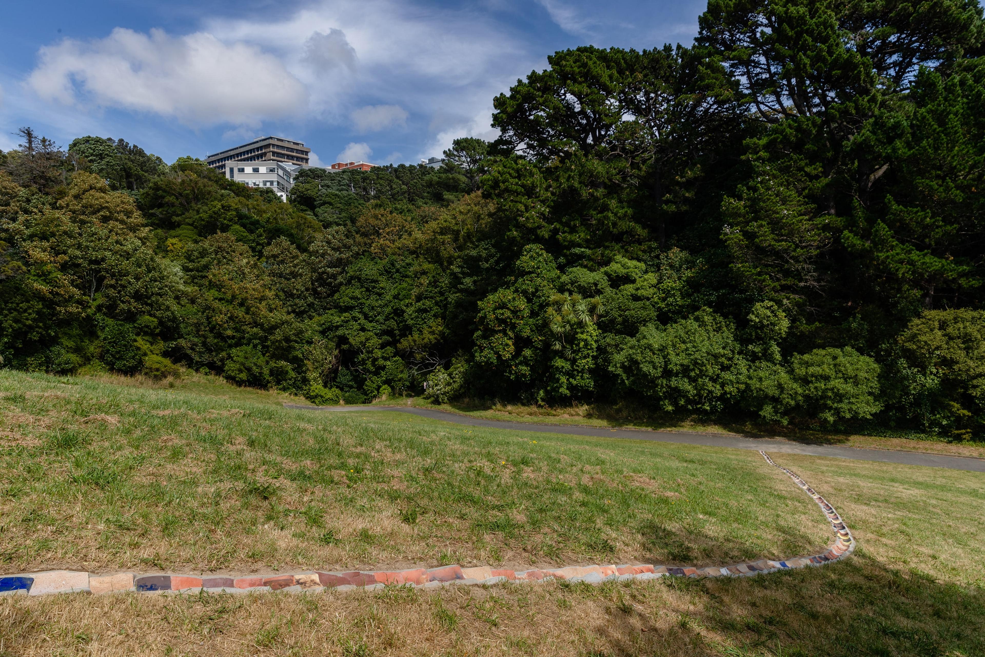 a long curving drain in a grass park, set with rounded terracotta tiles of different colours. There are large trees and a building on a hill in the background