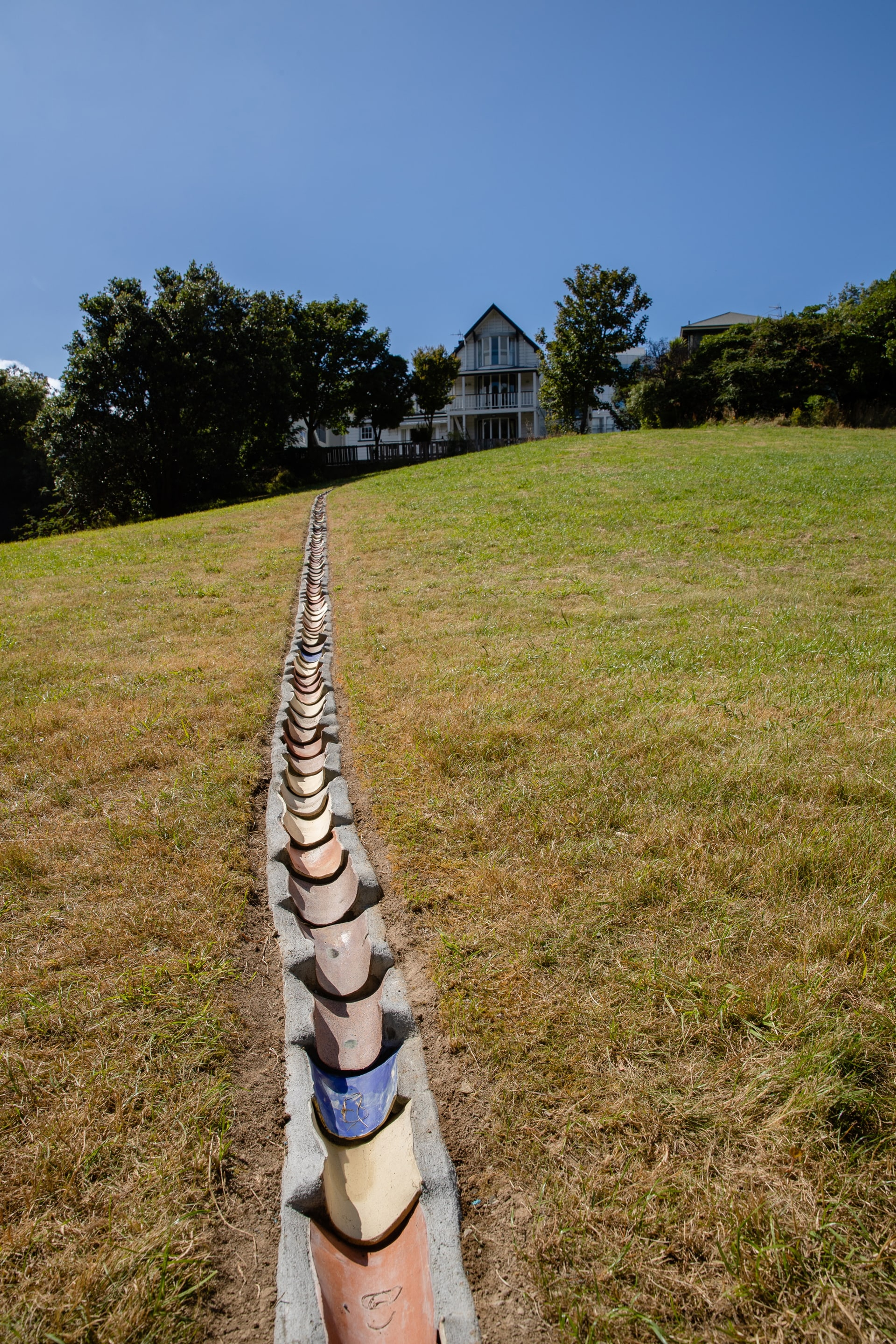a long drain in a grass park, set with rounded terracotta tiles of different colours. There is blue sky and a house in the background