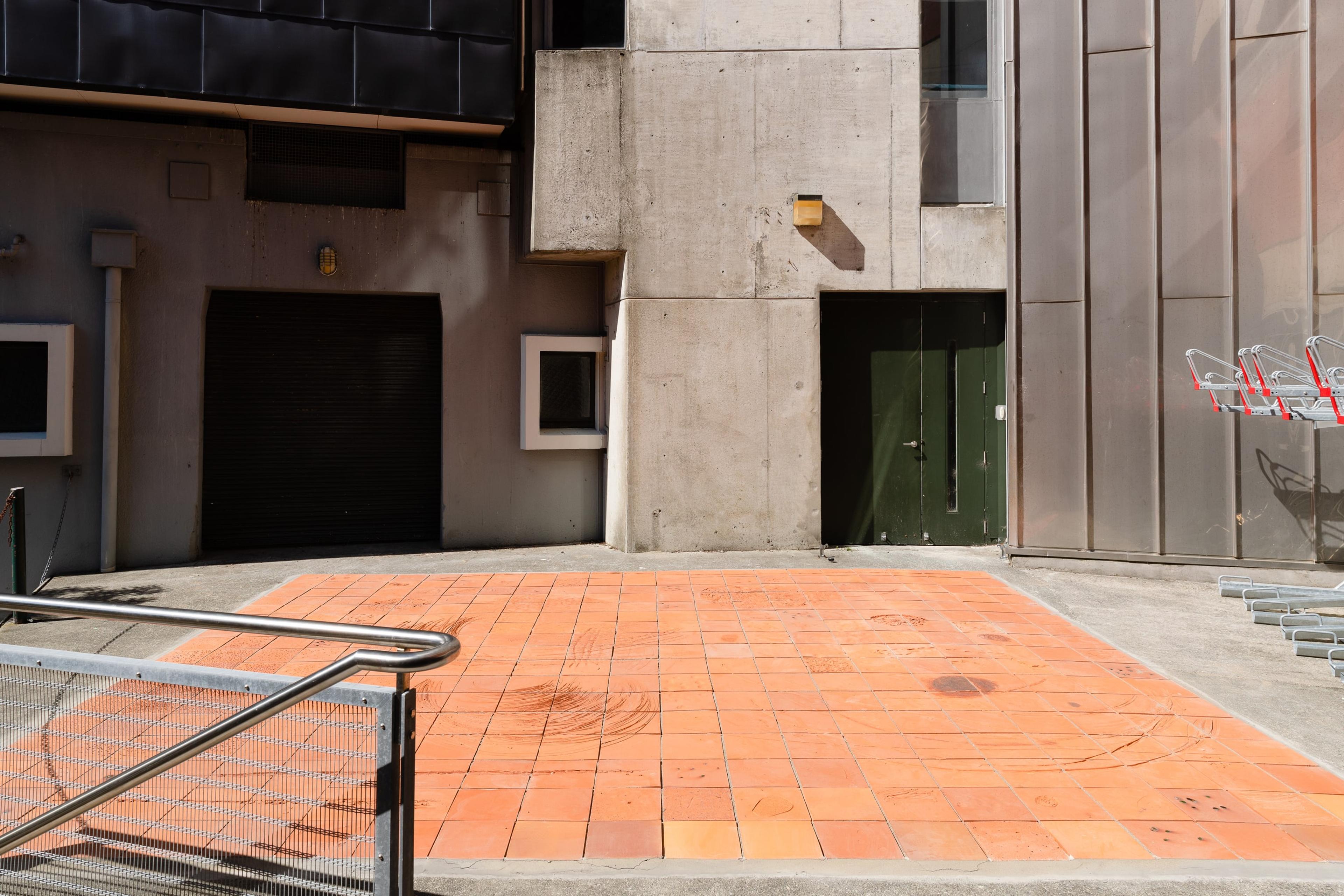 photograph of a flat area of terracotta tiles next to a grey concrete building and a stair rail obscuring it on the left side