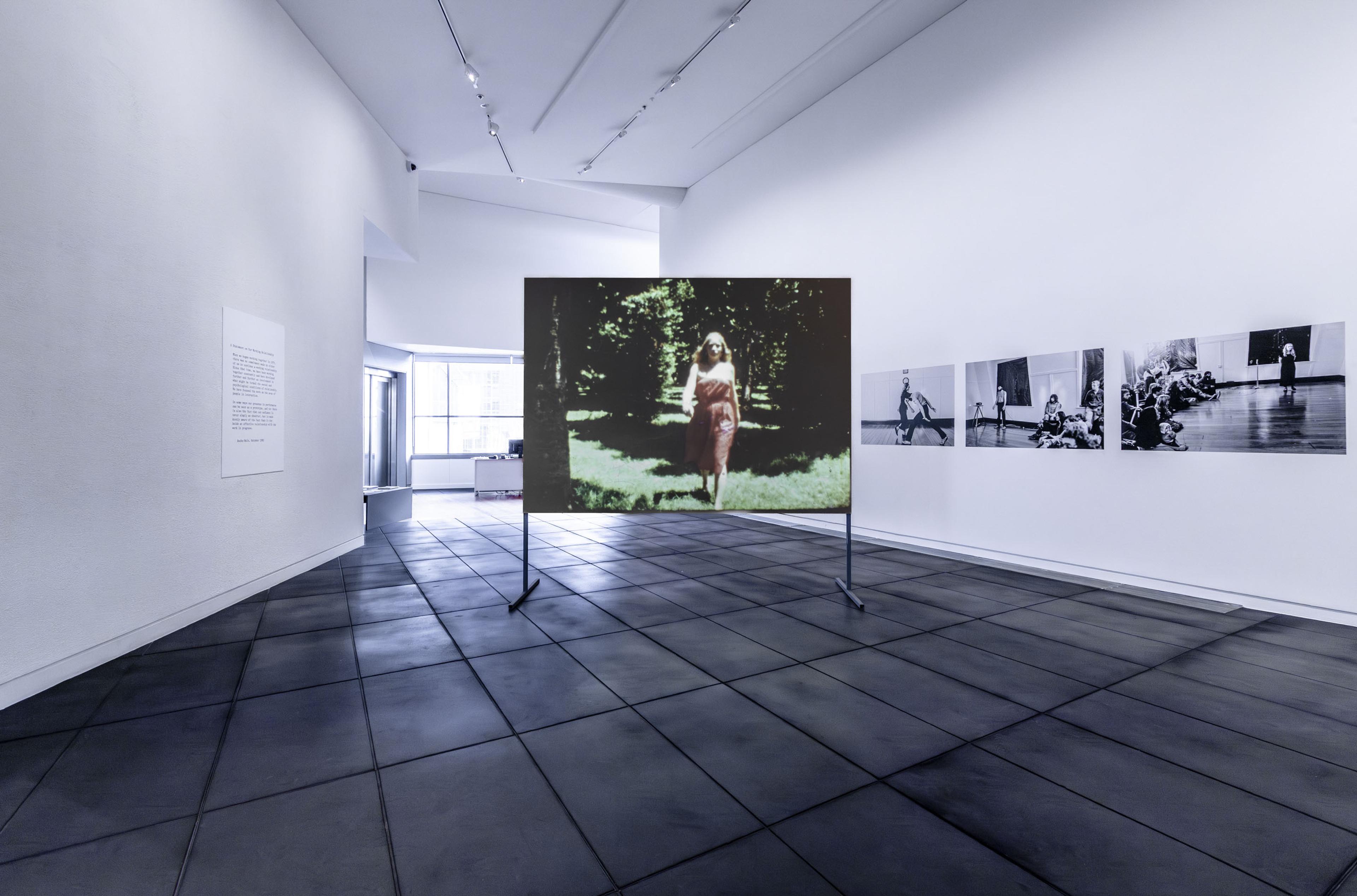 Installation image of the exhibition In Relation, a screen in the centre of the gallery's foyer with a film showing a woman in a red dress (Linda Buis) walking towards the camera. Black and white photographs of other performances are on the foyer wall.