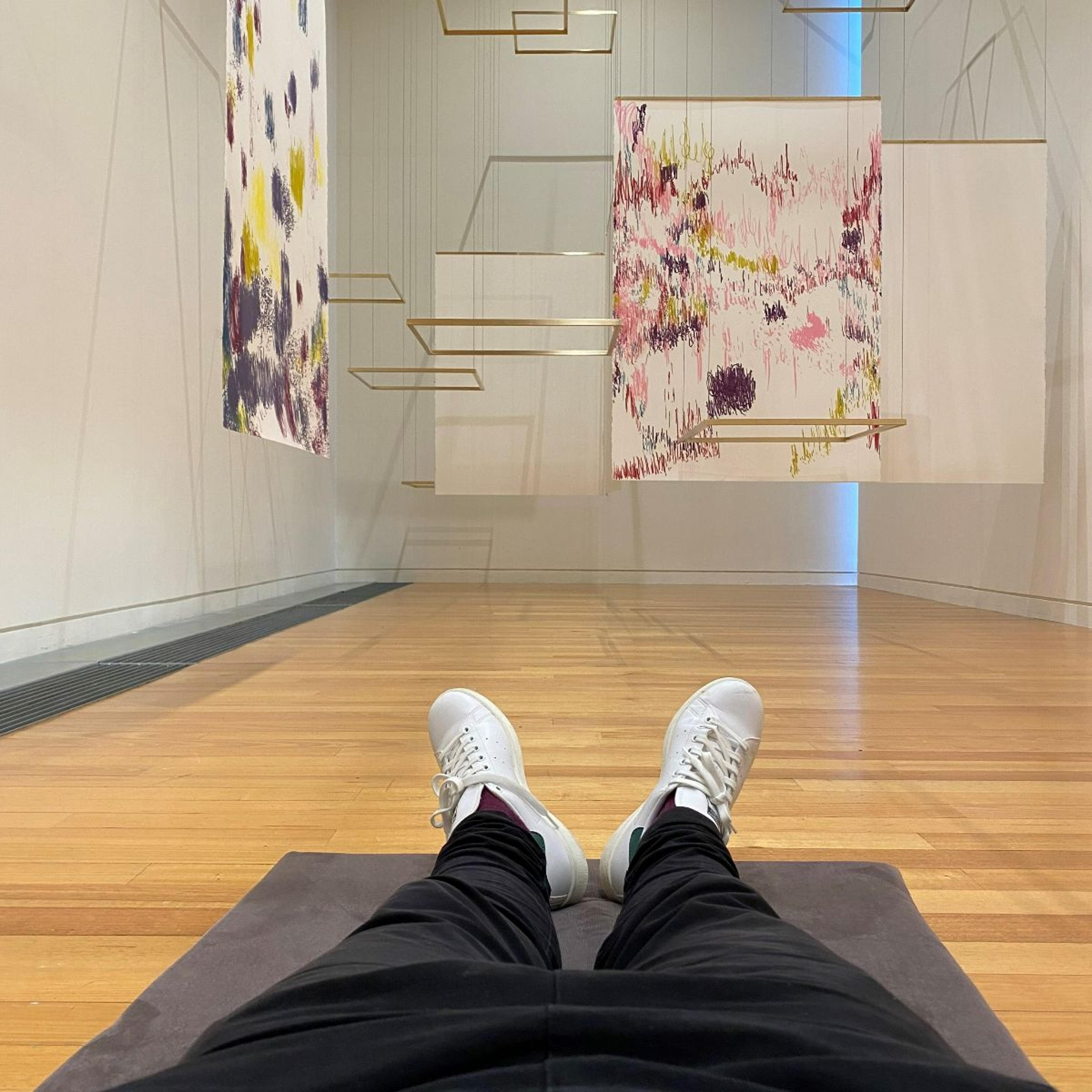 Person laying down on the floor underneath colourful drawings and brass frames hanging above