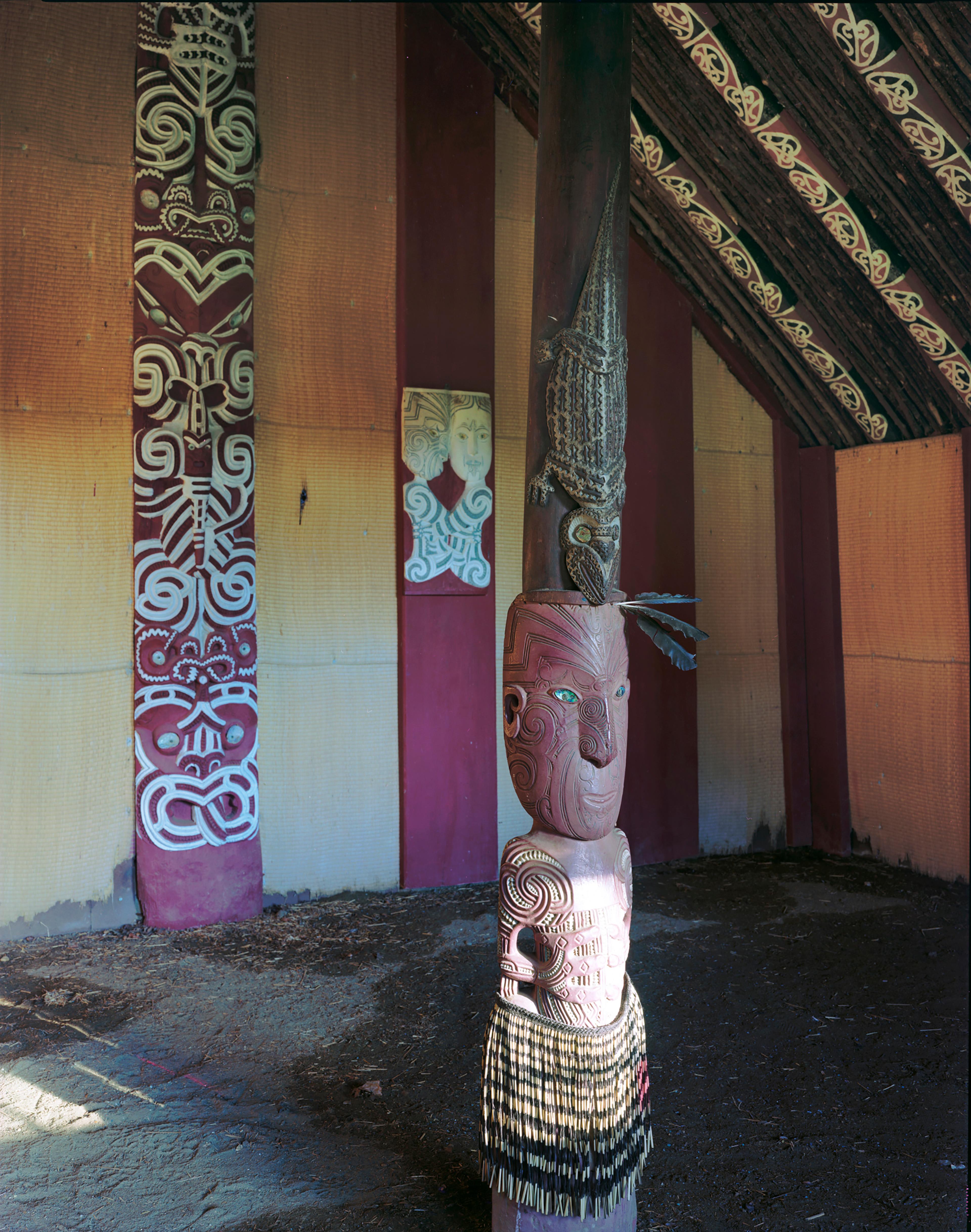 Image of carved pou at entrance to Hinemihi with carved and painted panels in back ground
