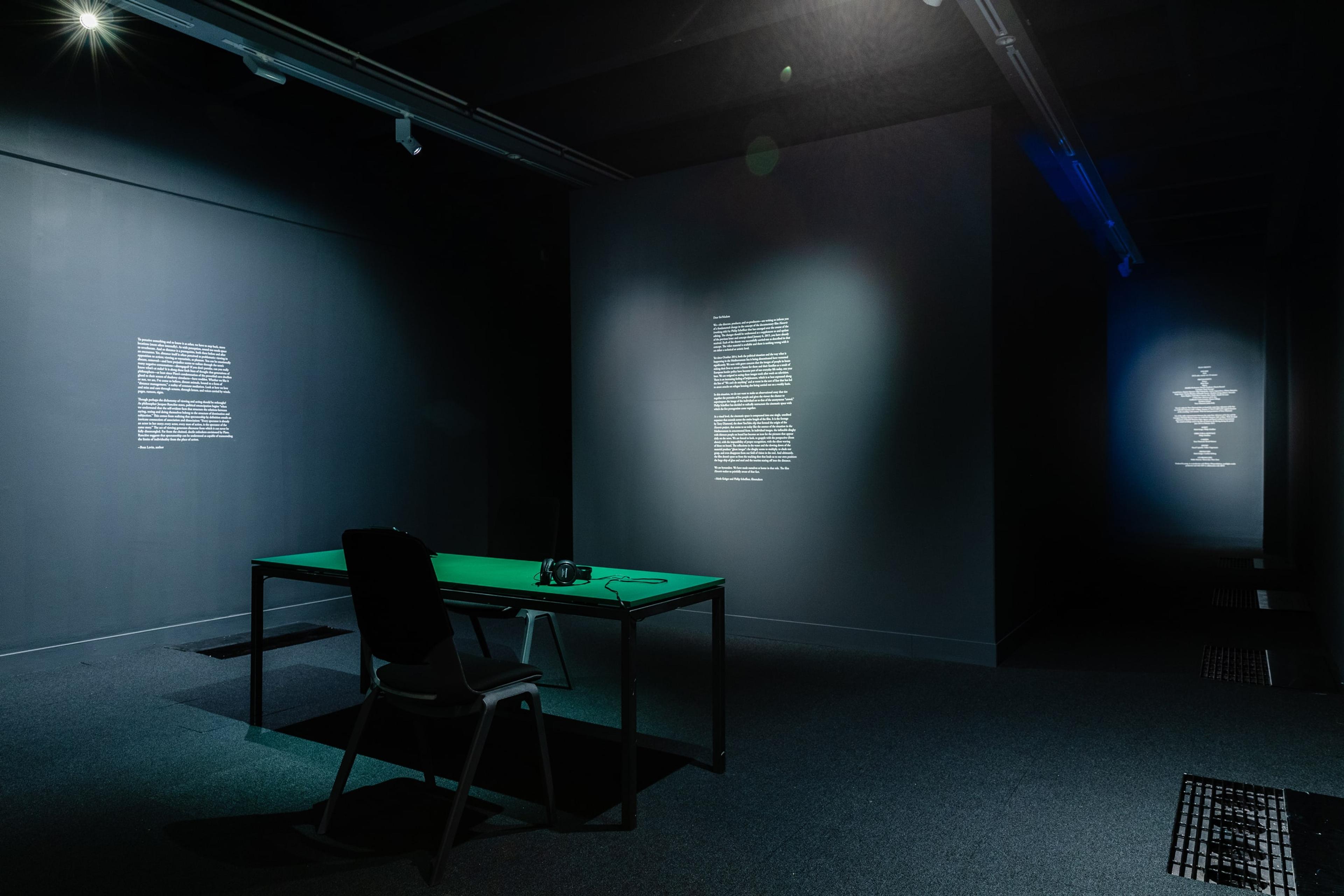 a dark room with black walls, with blocks of small white text spot-lit on the walls. A desk and chair sit in the middle of the room.