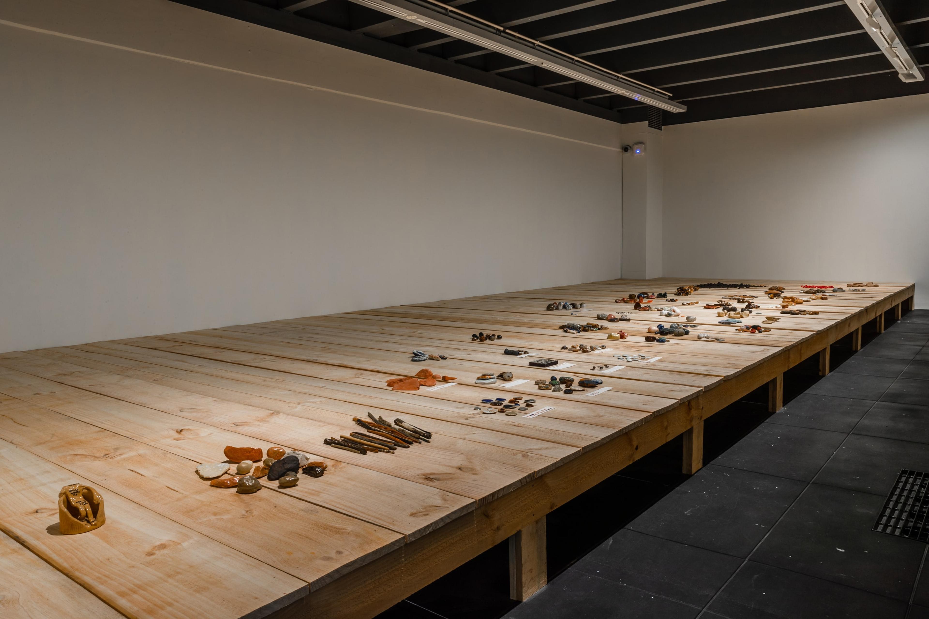 A wooden stage in a dimly lit white walled gallery, covered with groups of small objects
