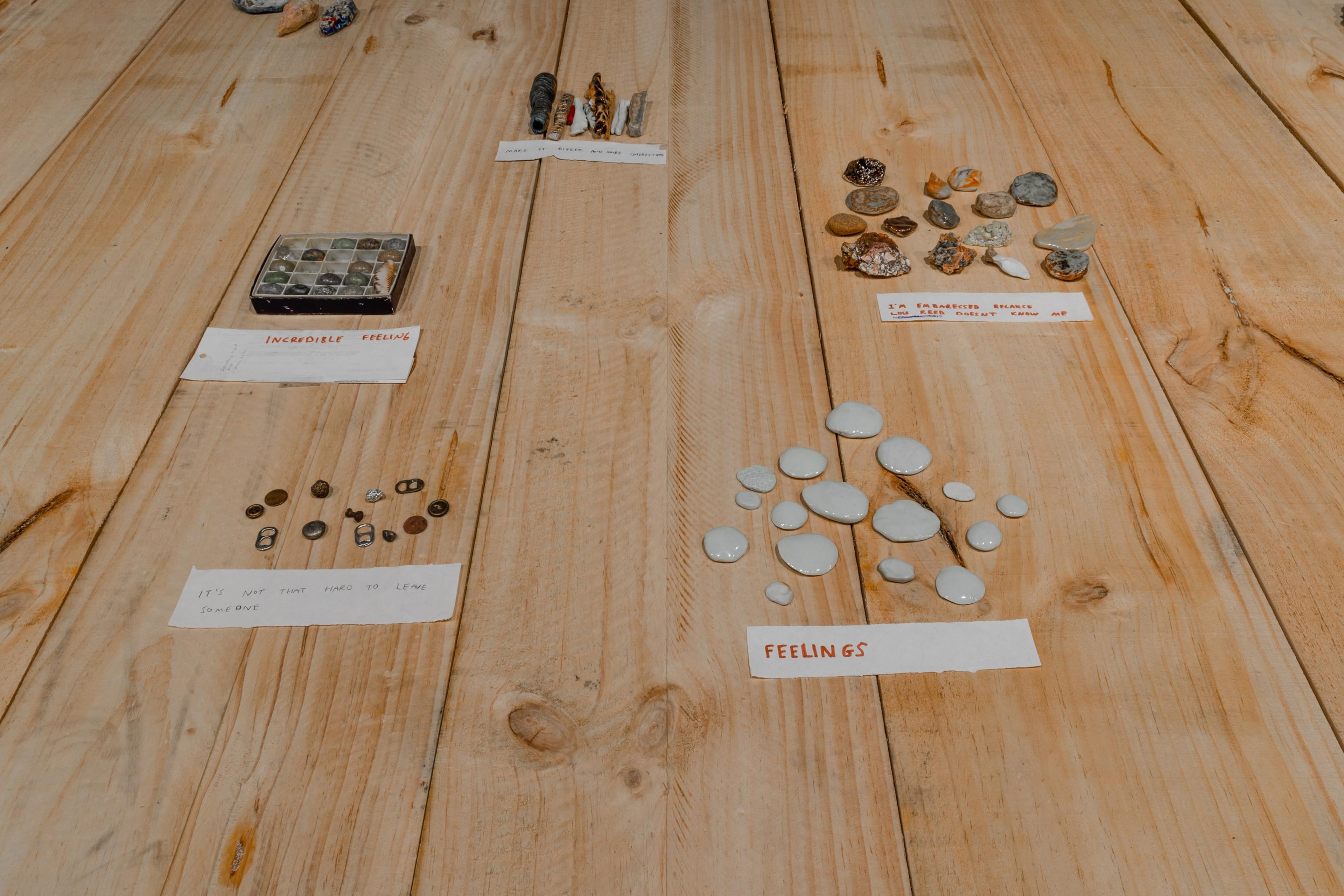 A wooden floor in a dimly lit gallery, covered with groups of small objects with written paper labels in red ink