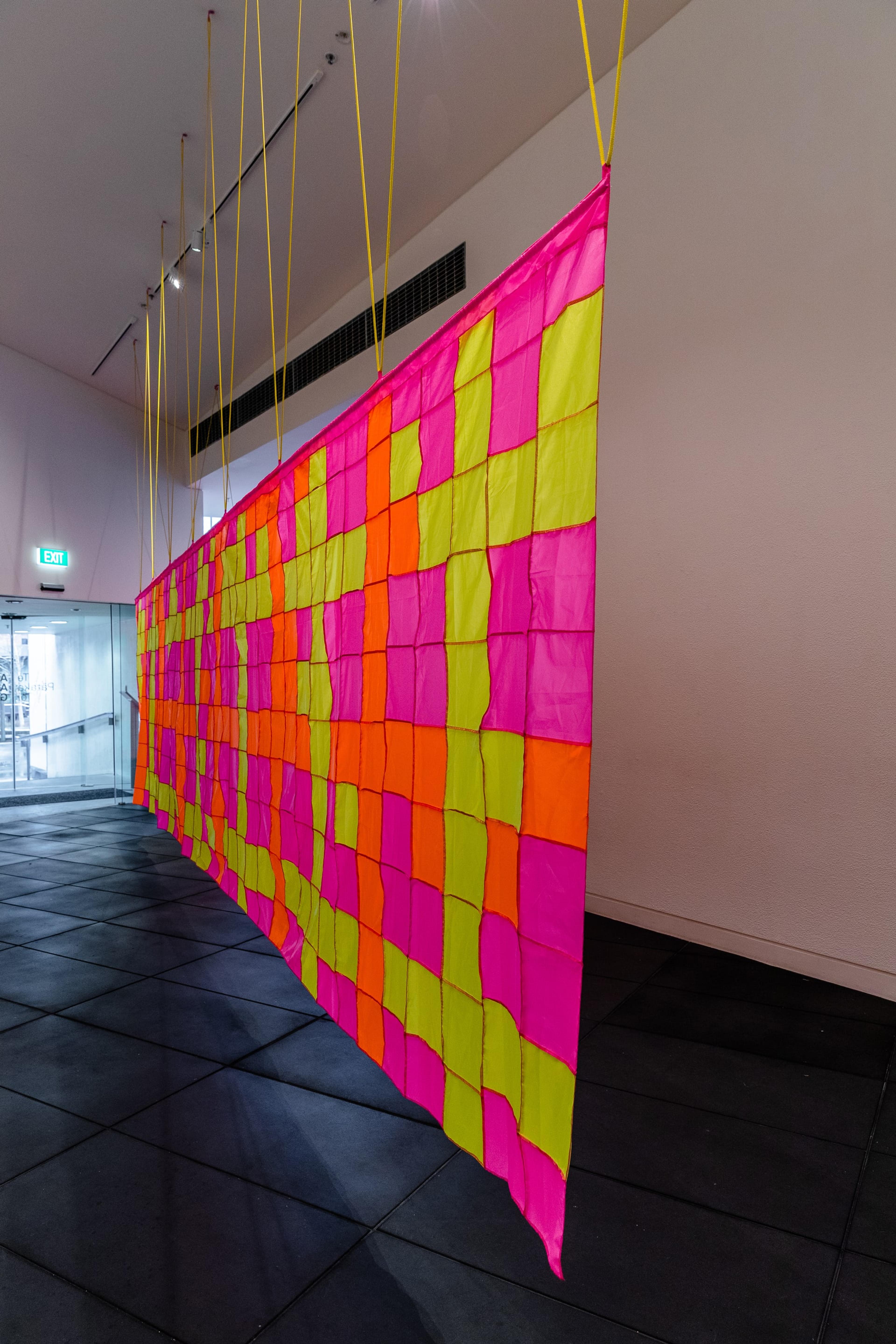 A large patchwork banner of fluoro yellow, pink and orange squares, hanging in a white walled, black floored gallery