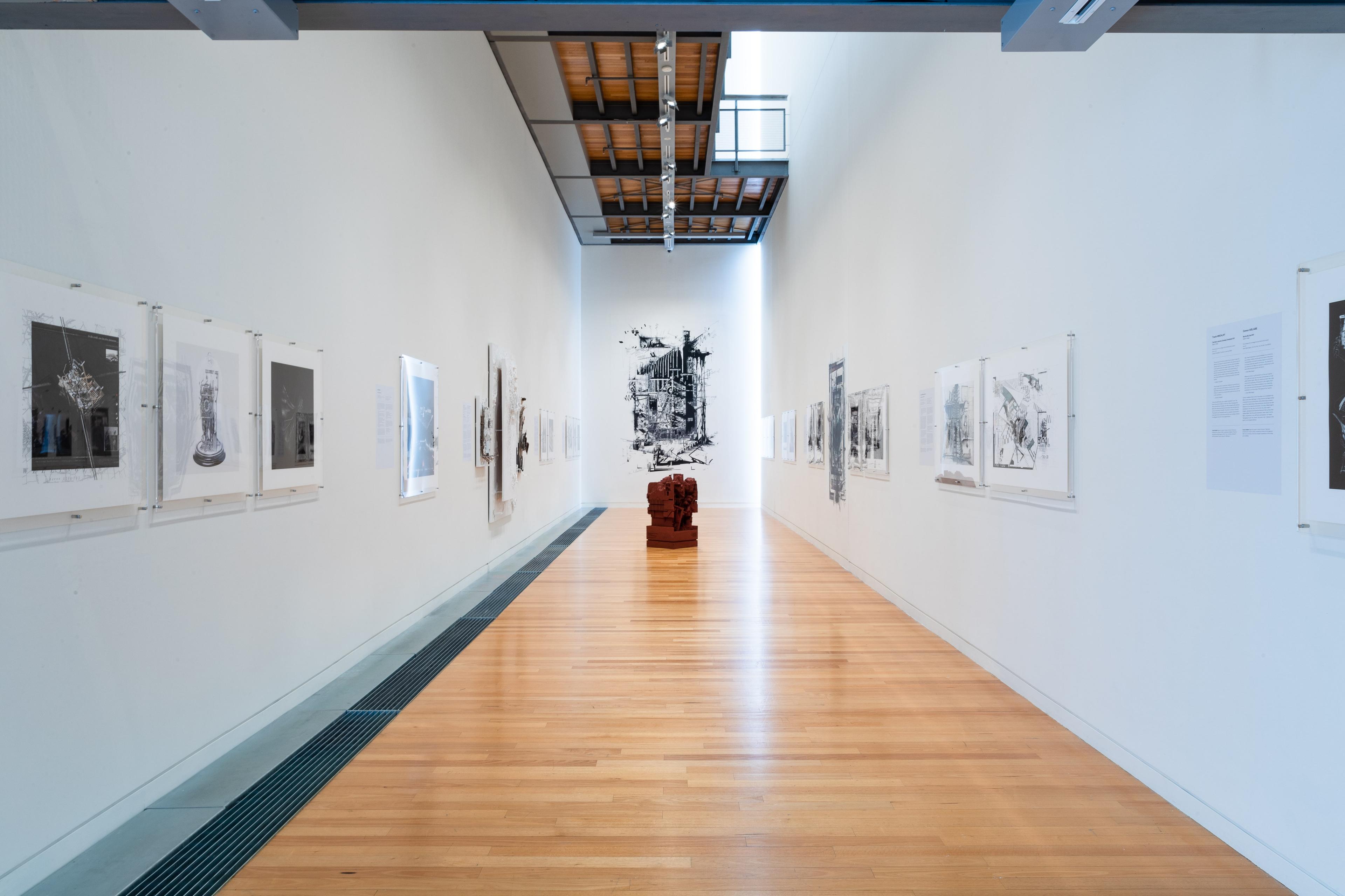 Wide view of a long gallery with text and artworks on the white walls and a dark red sculpture on the wooden floor. Daniel K. Brown 