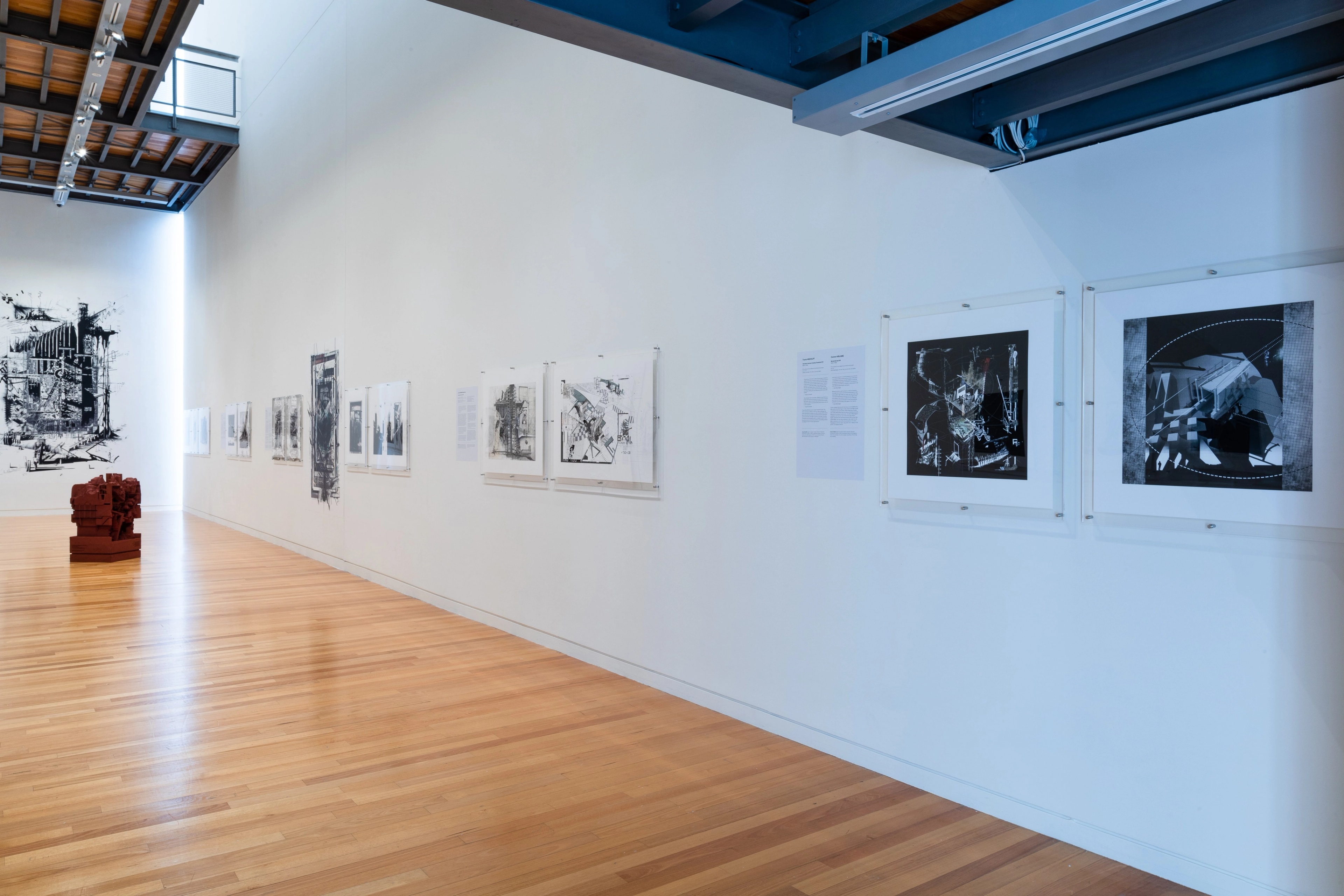 Wide view of a gallery with text and artworks on the white walls and a dark red sculpture on the wooden floor. Daniel K. Brown 