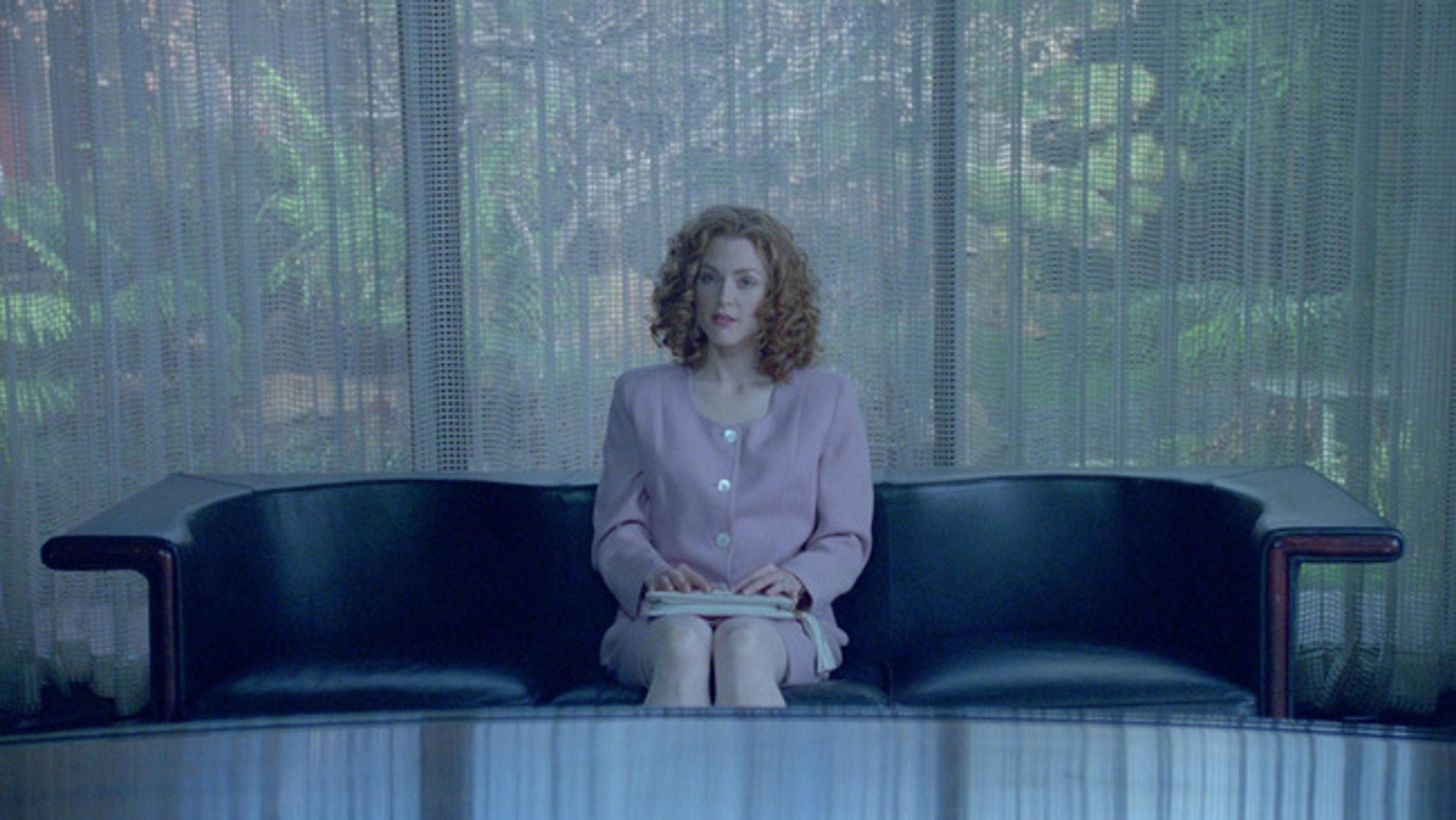 Still image women sitting on leather couch in pink suit, exterior view of garden through thin curtains