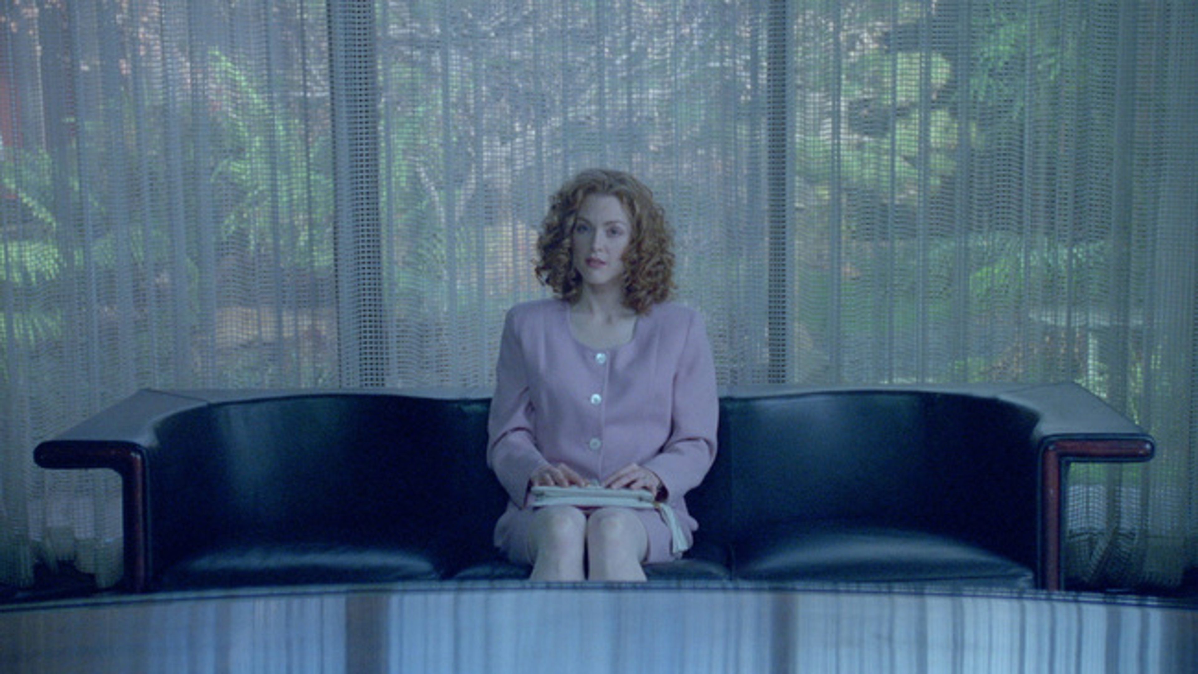 Still image women sitting on leather couch in pink suit, exterior view of garden through thin curtains