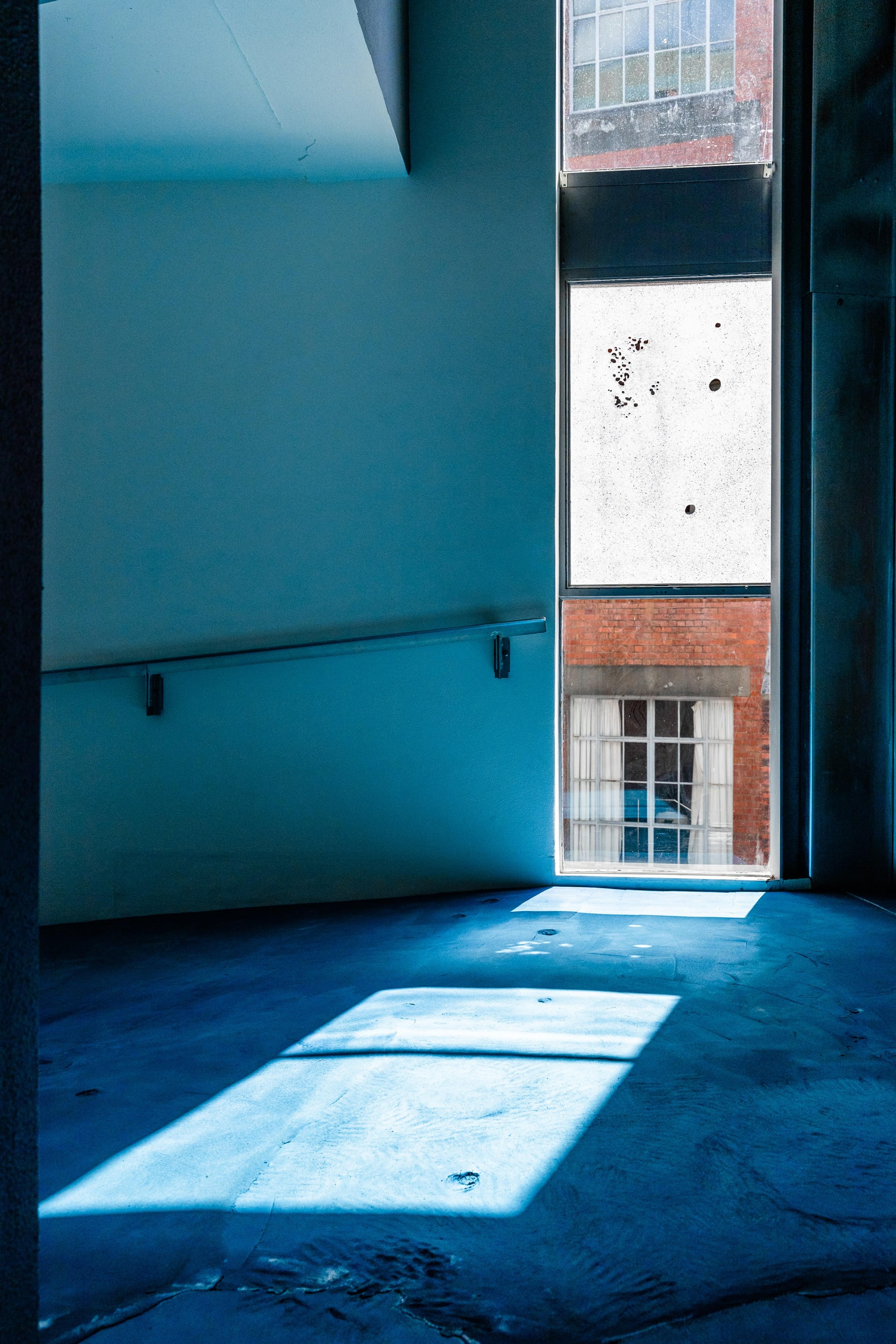 A view of the textured blue ground inside the gallery, with sunlight streaming in and blue light reflecting up onto the white walls. Outside the window is brick building, and one of the windows is frosted glass with several round holes in it