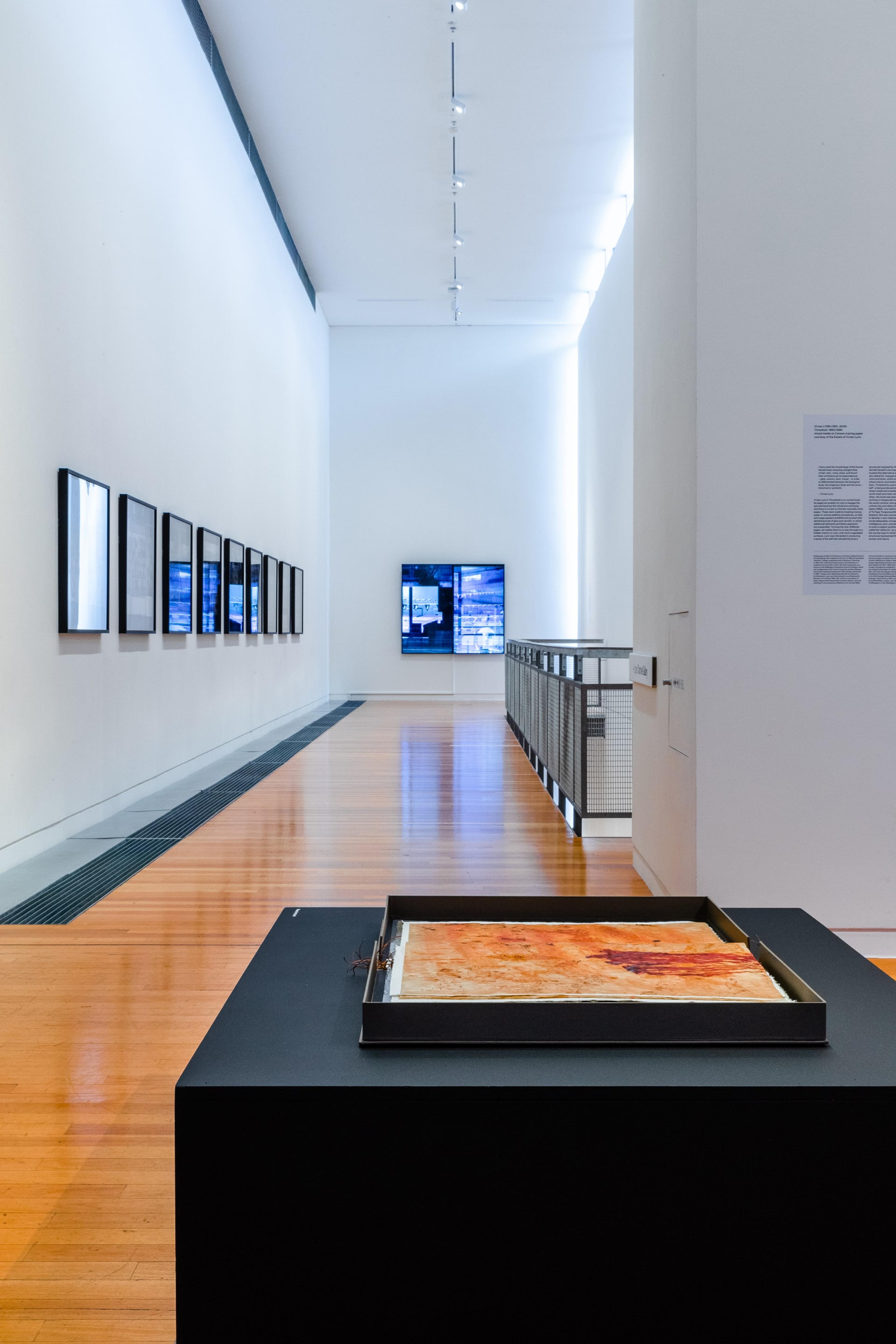 A wider view of the gallery, with a flat brown and white artwork on a low black bench table in the foreground and images on the walls.