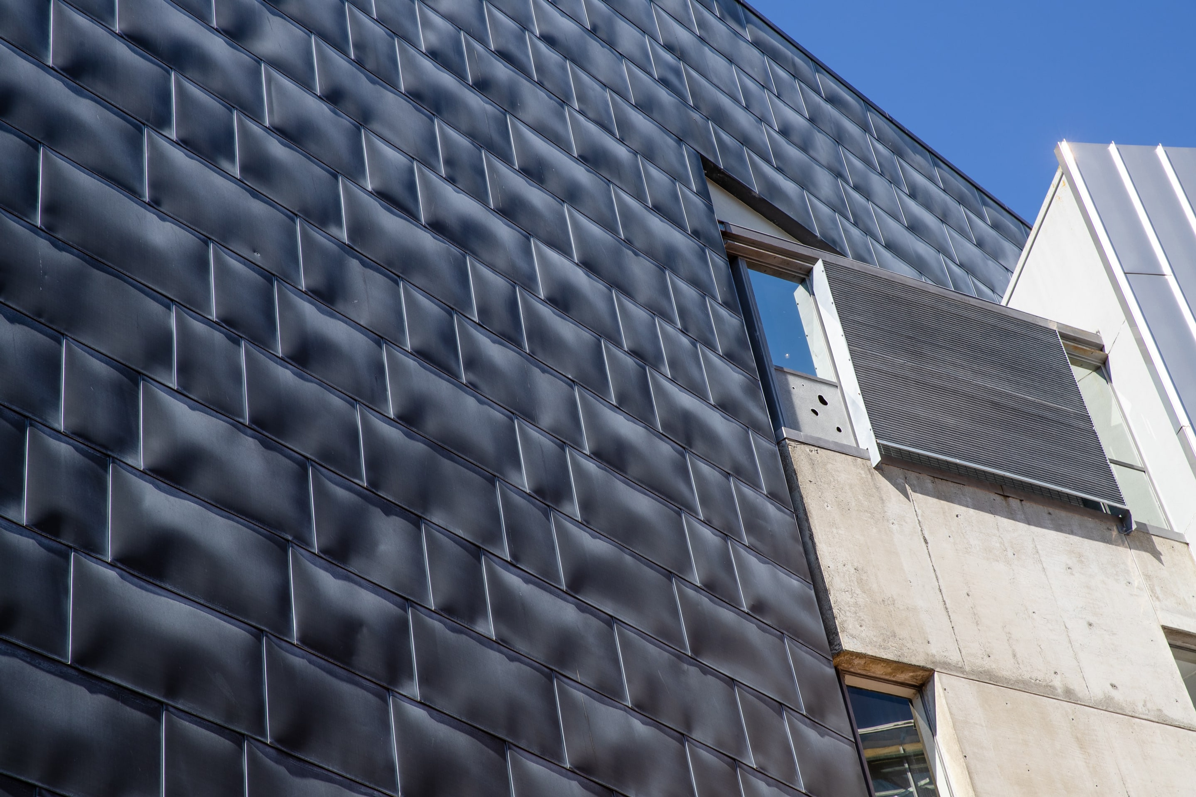 Photo of the outside rear of the gallery, looking up at a grey tiled wall and an angled grate and frosted window with several small holes