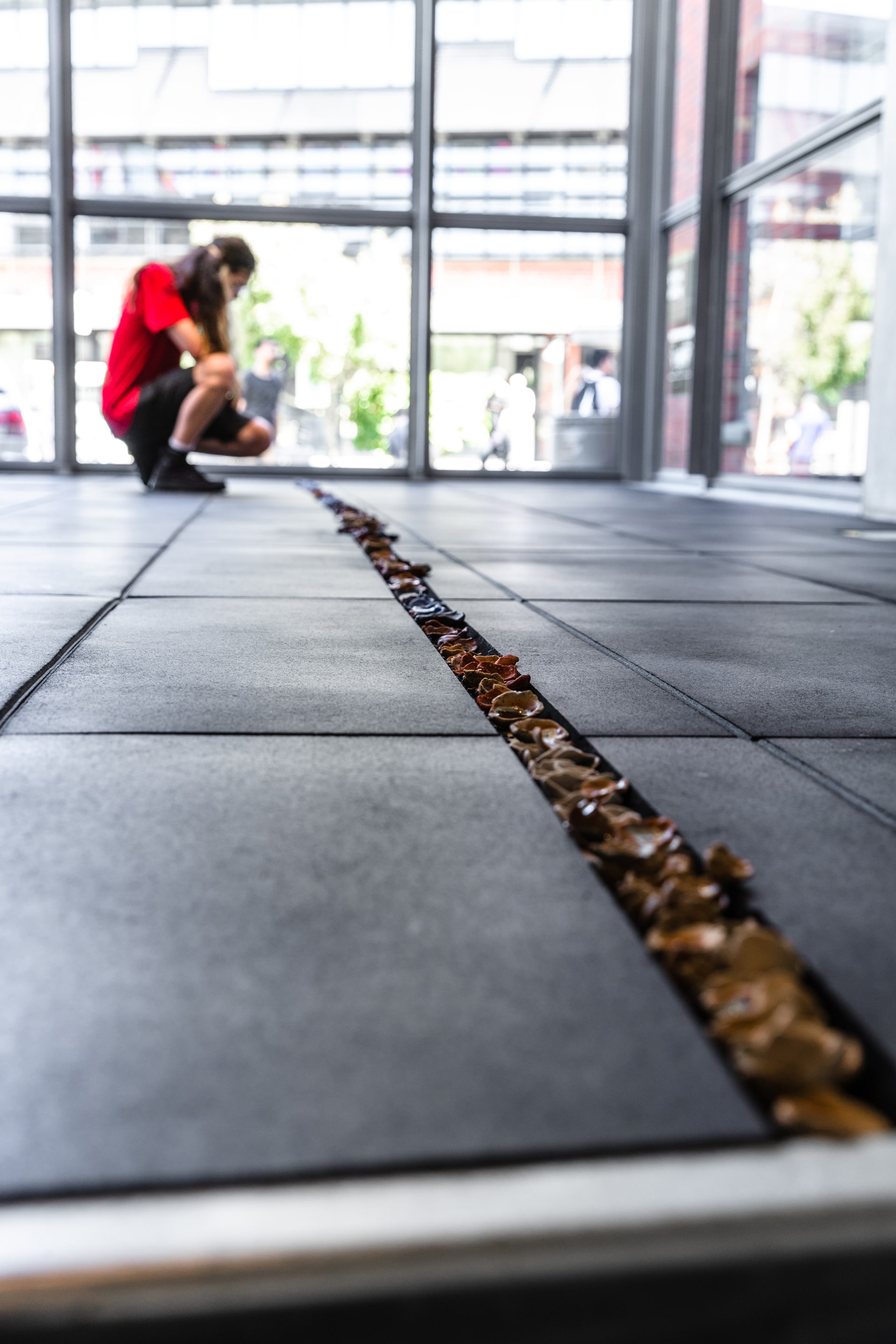 a floor level view of groove in rubber flooring tiles filled sith small ceramic objects. a person is crouching at the end of the groove in front of the windows
