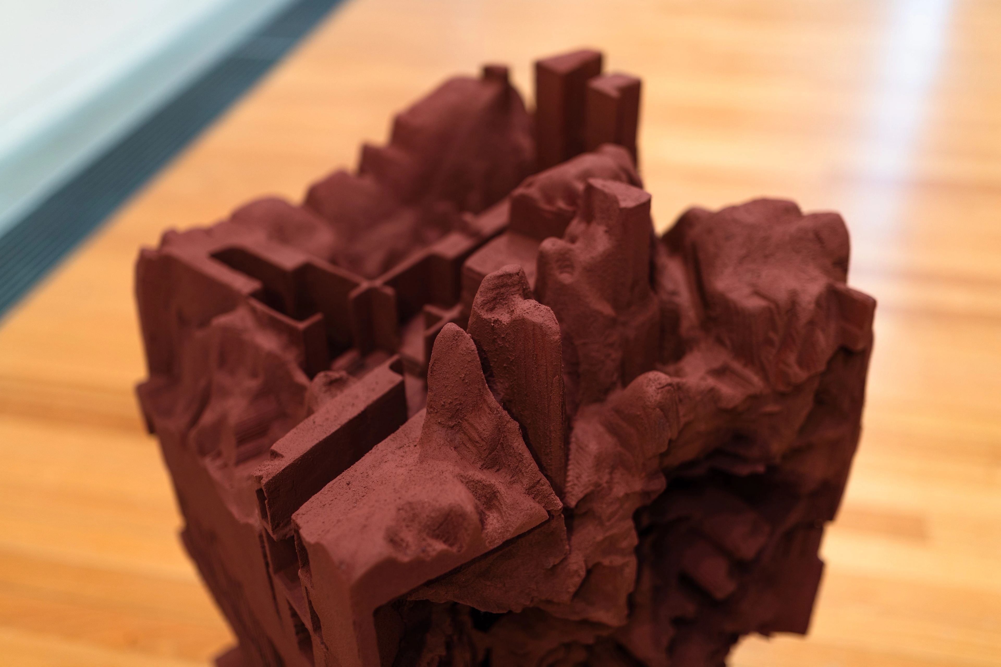 A wooden gallery floor with a dark red architectural sculpture, with geological looking lumps and square angles