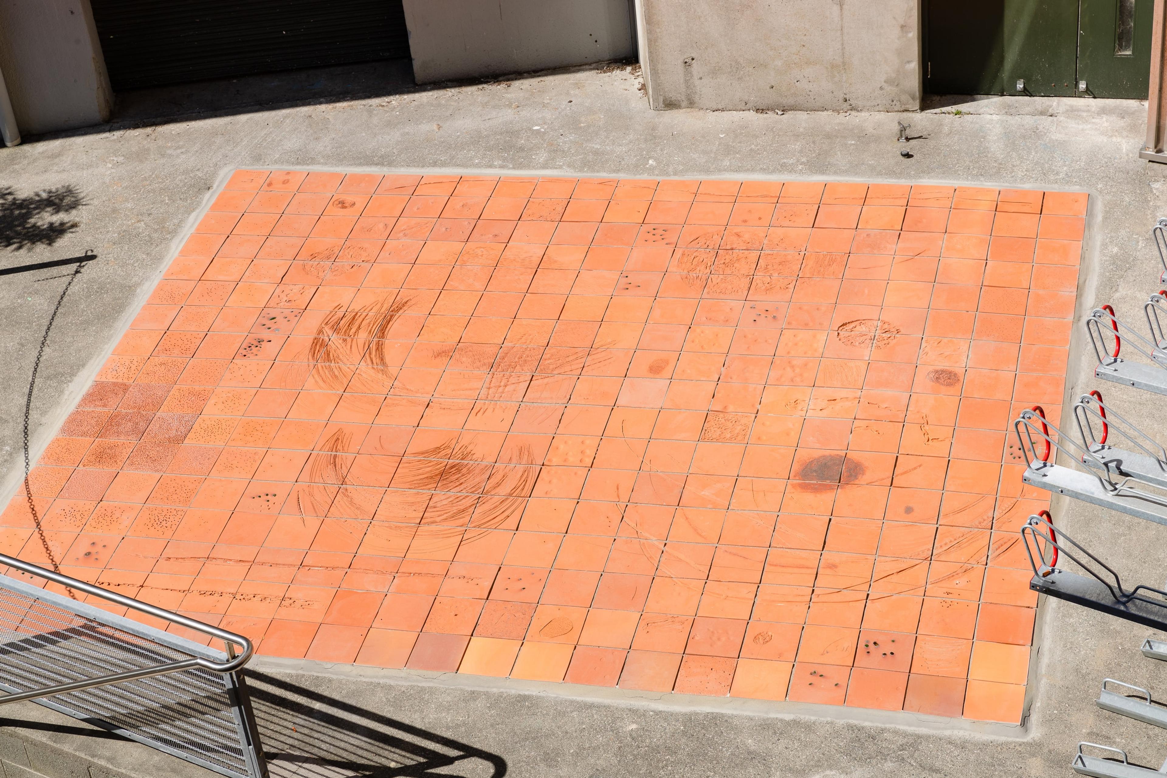 an outdoor flat tiled area covered in terracotta tiles with large scratches and marks, and metal bike racks and a stair railling on either side