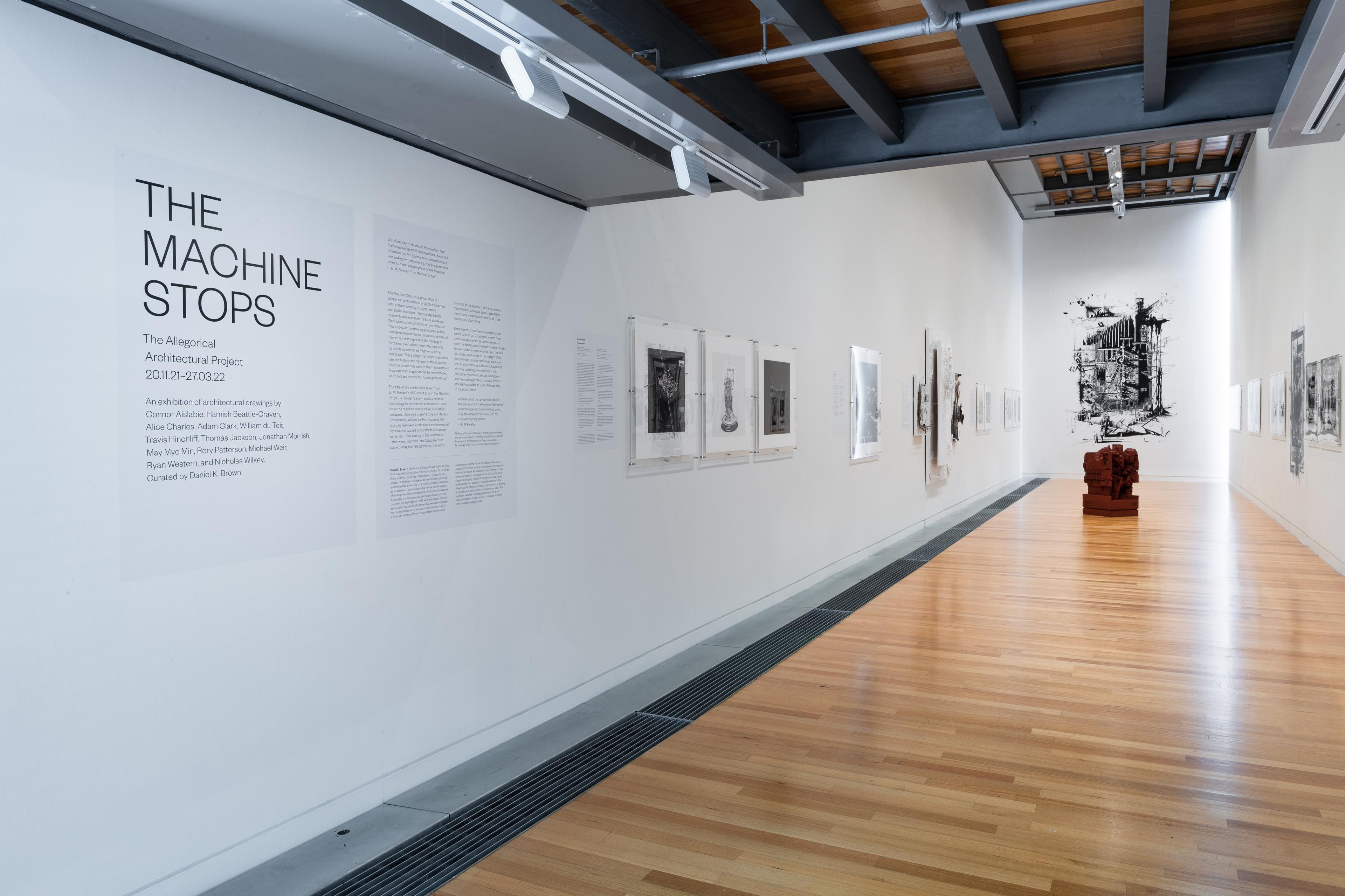Wide view of a gallery with text and artworks on the white walls and a dark red sculpture on the wooden floor. Daniel K. Brown 