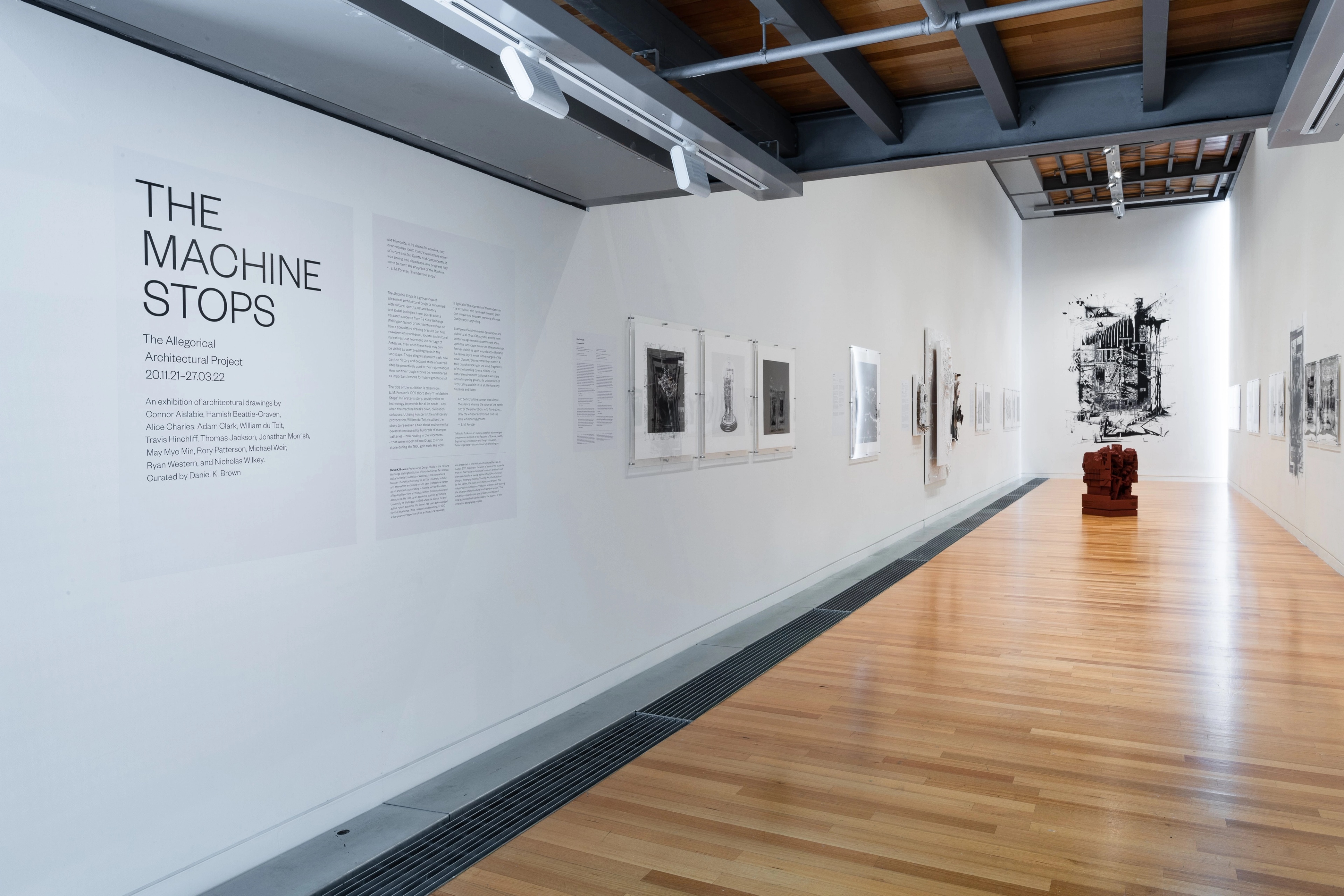 Wide view of a gallery with text and artworks on the white walls and a dark red sculpture on the wooden floor. Daniel K. Brown