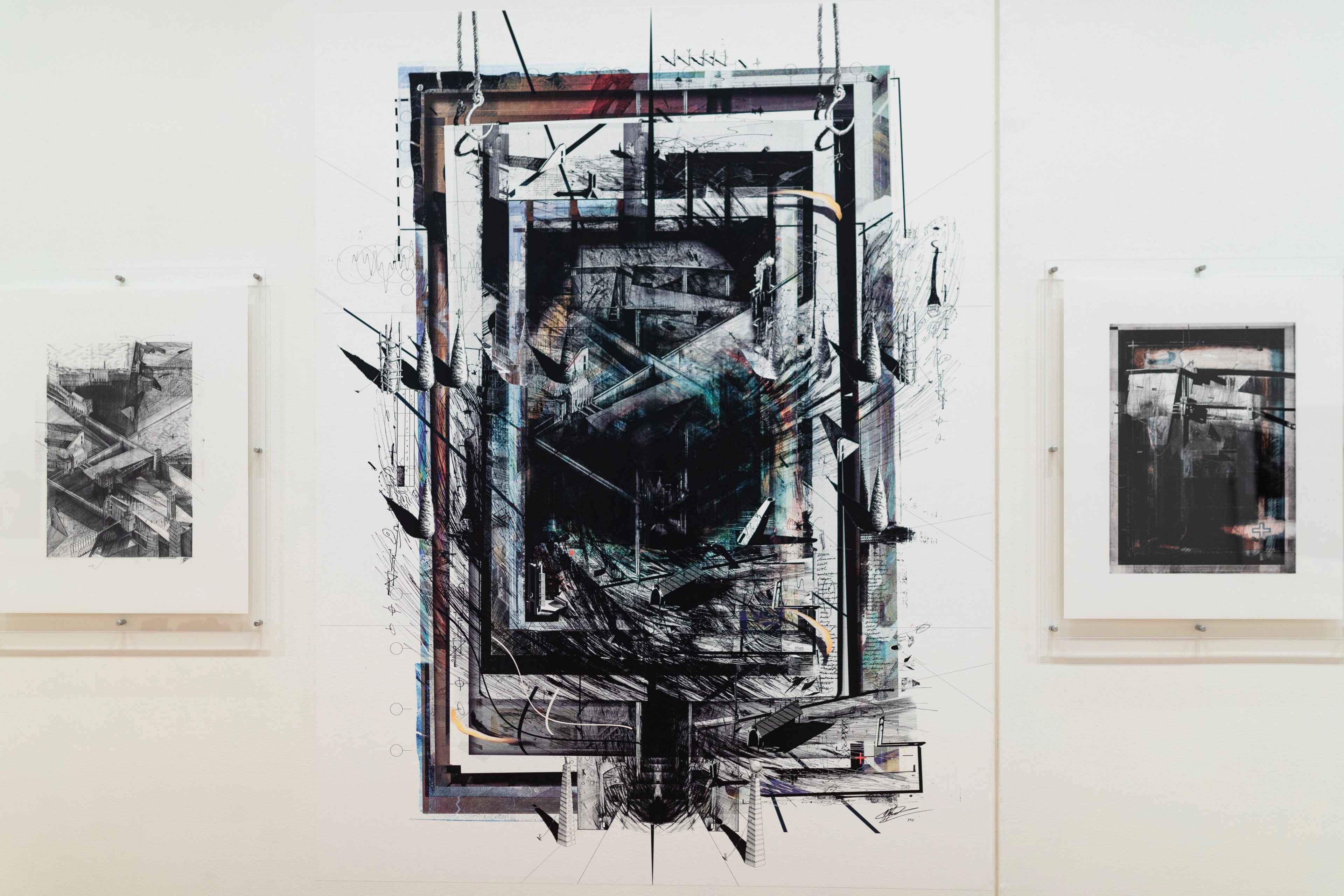 A straight on view of a white gallery wall with a square architectural image in perspex mount on either side of a larger print of a dark angular architectural drawing . Daniel K. Brown 