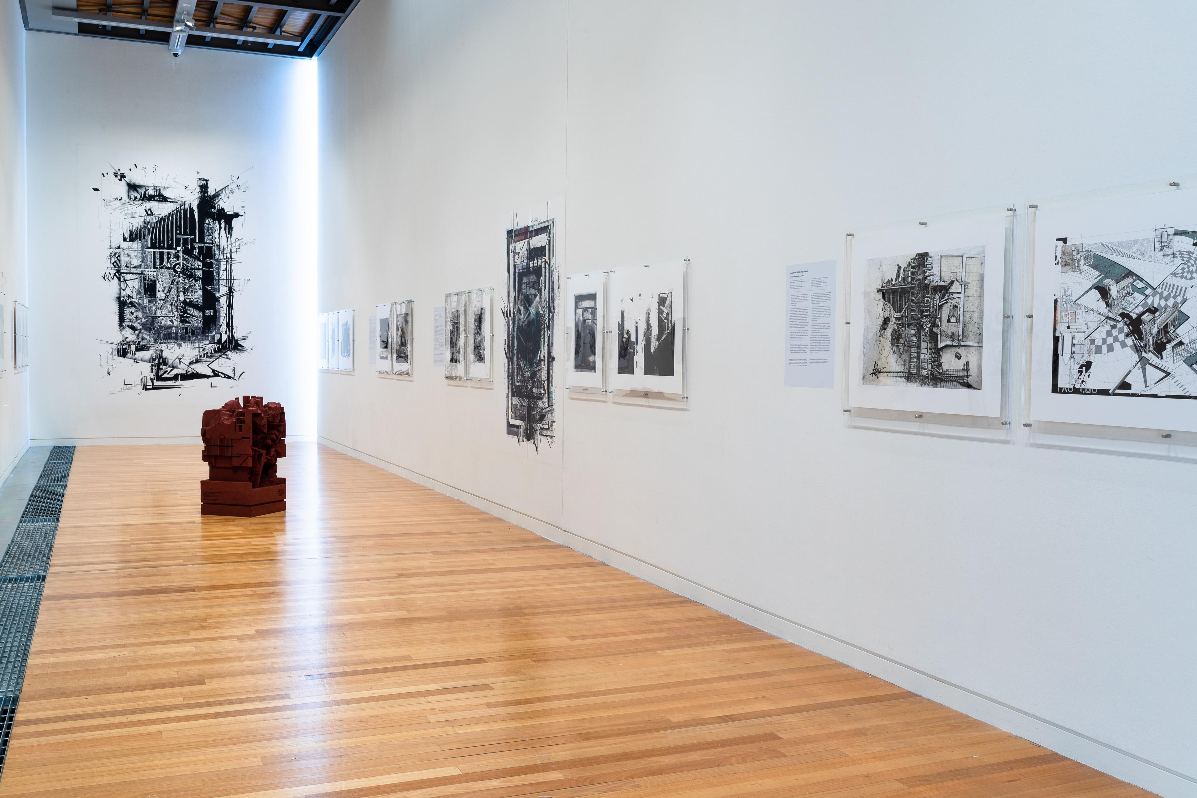 Wide view of a gallery with text and artworks on the white walls and a dark red sculpture on the wooden floor. Daniel K. Brown 
