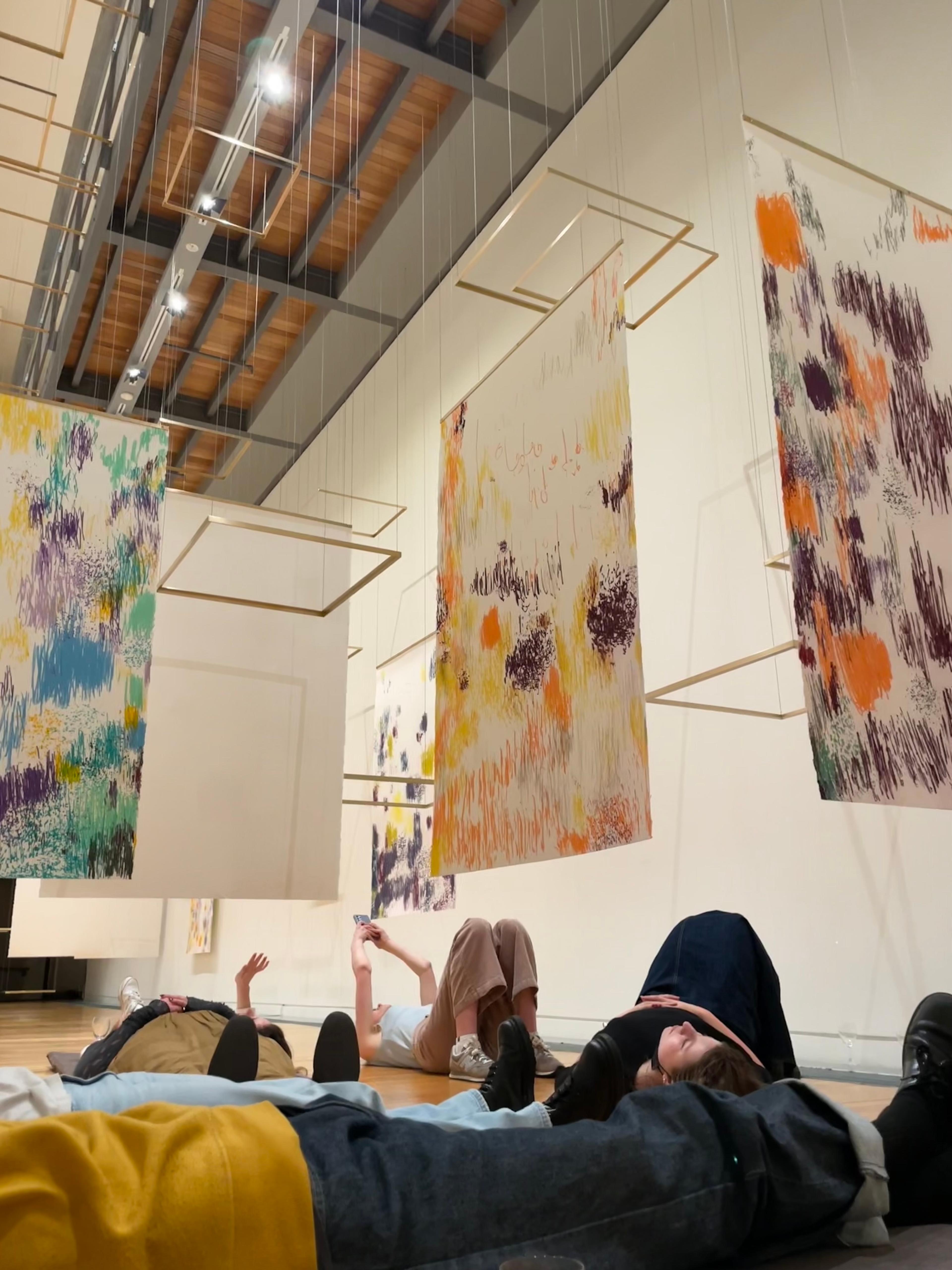 Adam Art Gallery volunteers laying on floor underneath hanging drawing and metal frames art work installation