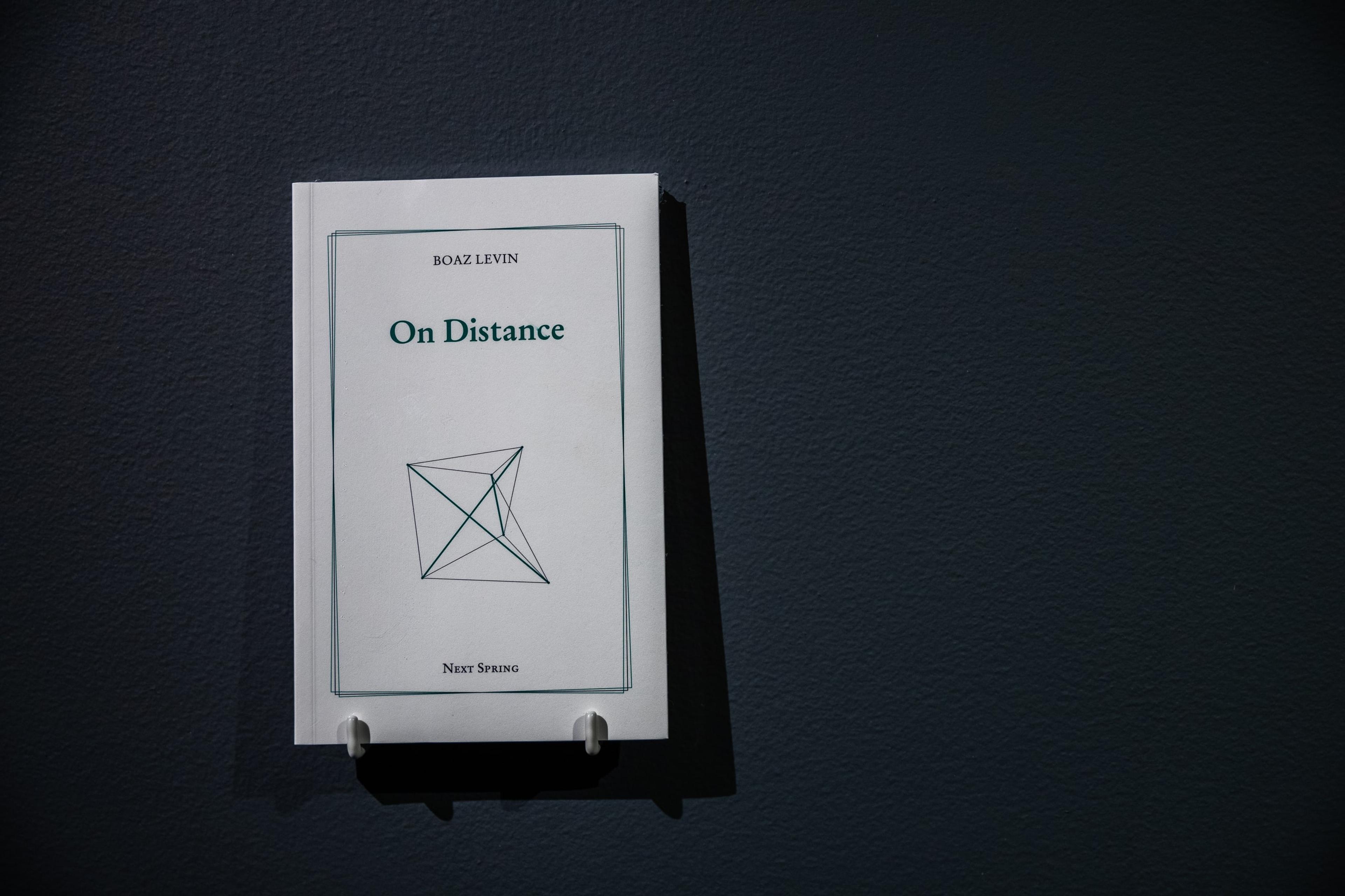 a black wall, with a small white paper booklet mounted on metal pegs. The booklet cover reads "on distance"