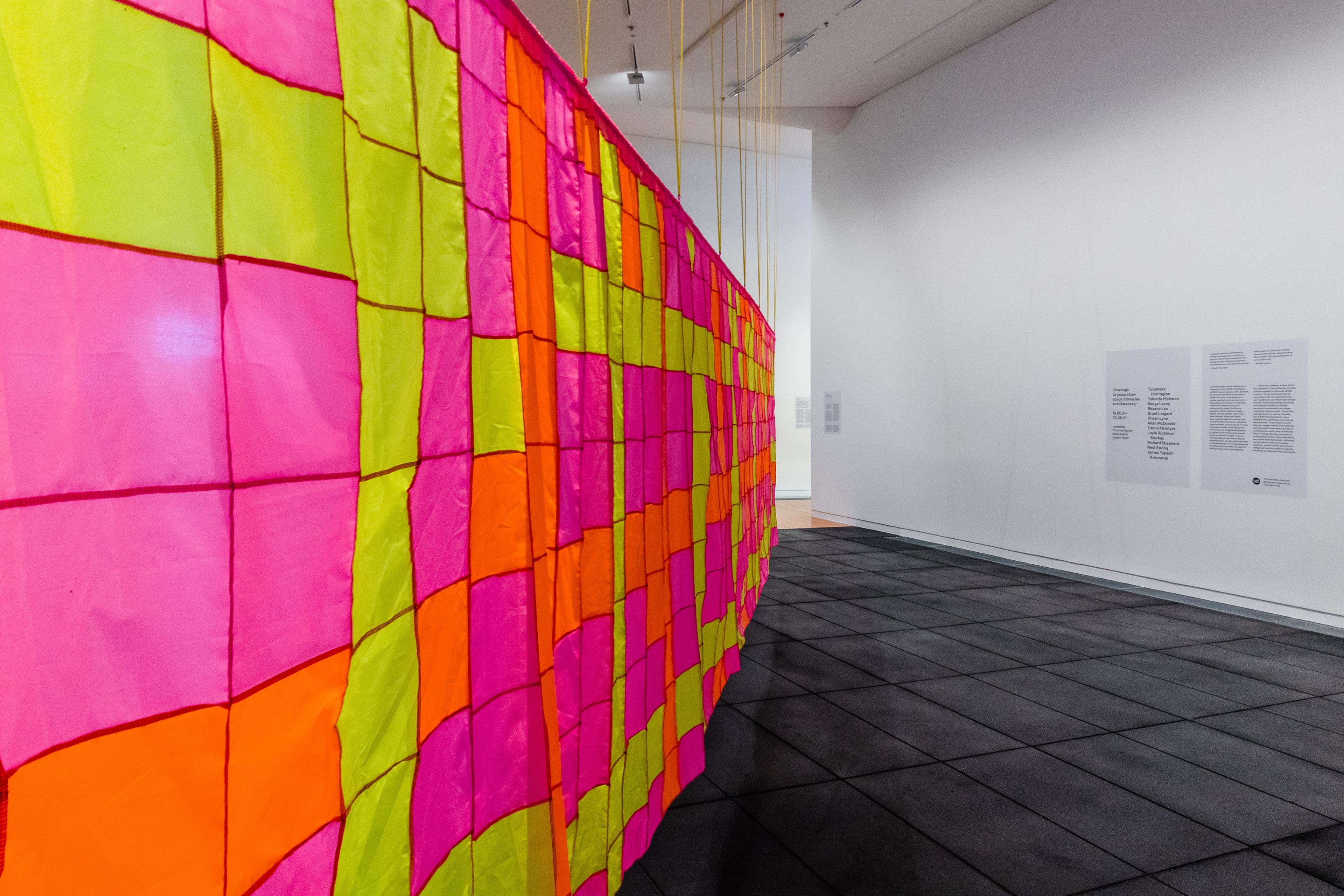 A large patchwork banner of fluoro yellow, pink and orange squares, hanging in a white walled, black floored gallery