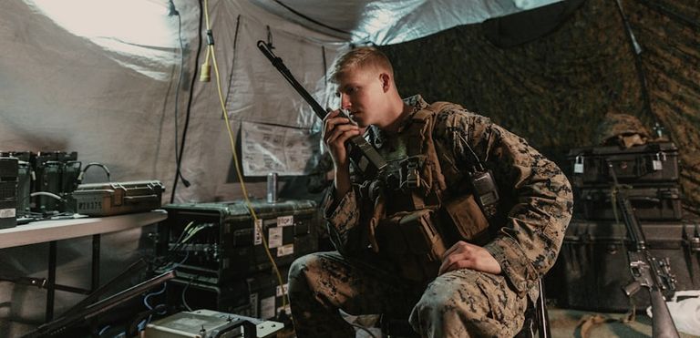 A soldier in fatigues speaks into a handheld radio inside a field communications tent.