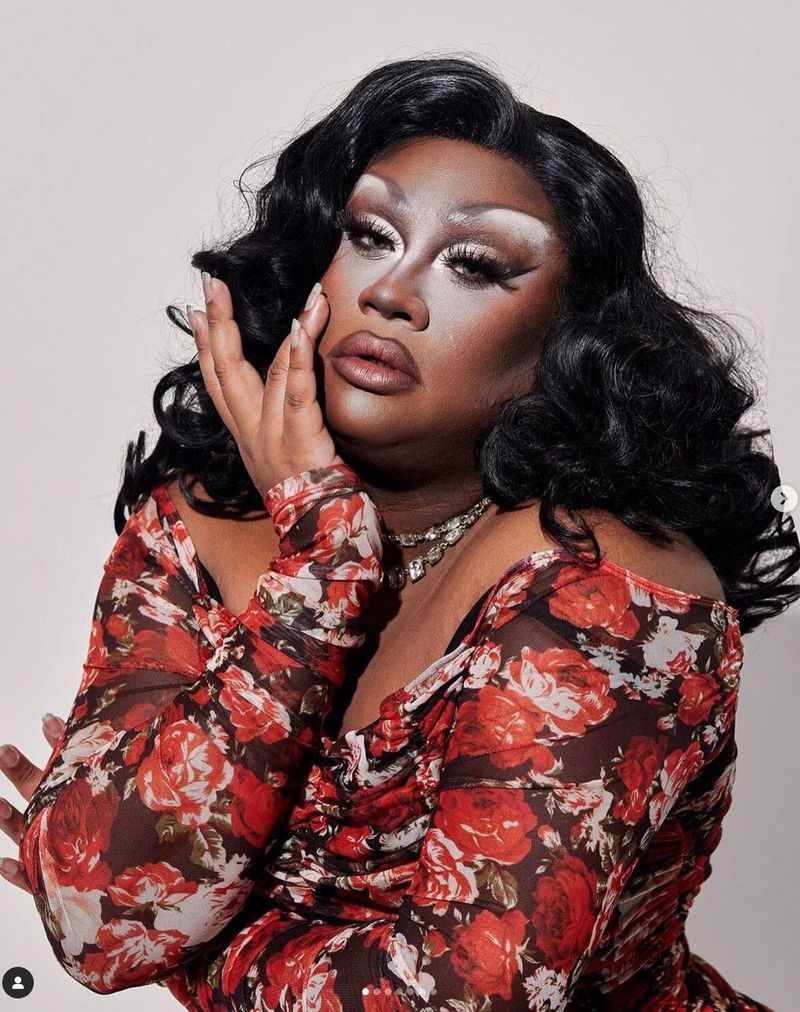 A closeup portrait of Mahatma posing in a red and brown floral top. She has black, long, glamorous Hollywood style hair.