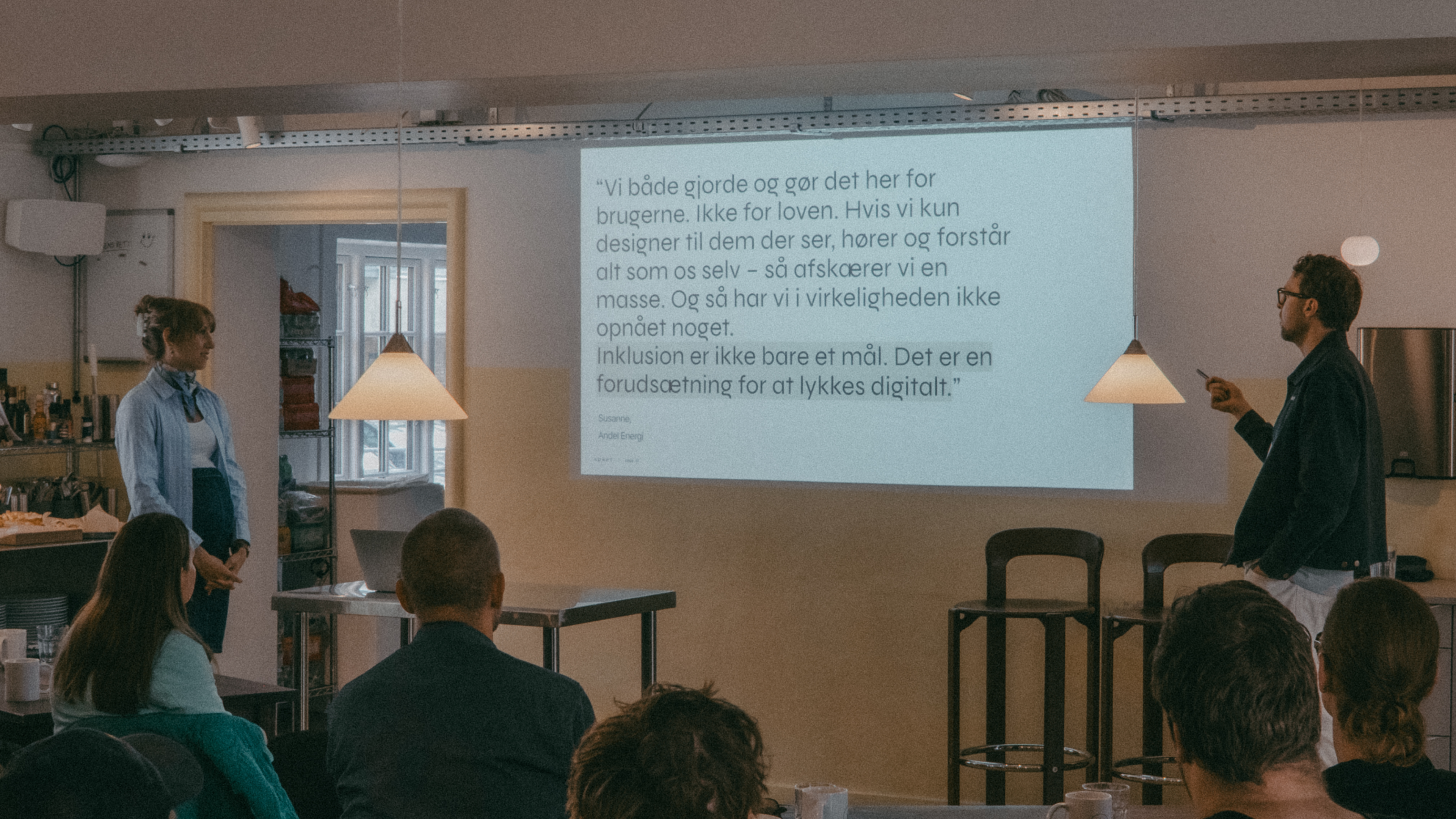 A woman and a man are giving a presentation in a bright room with a projecter, showing the presentation. The man is gesturing while speaking. Several people are seated and attentively listening.